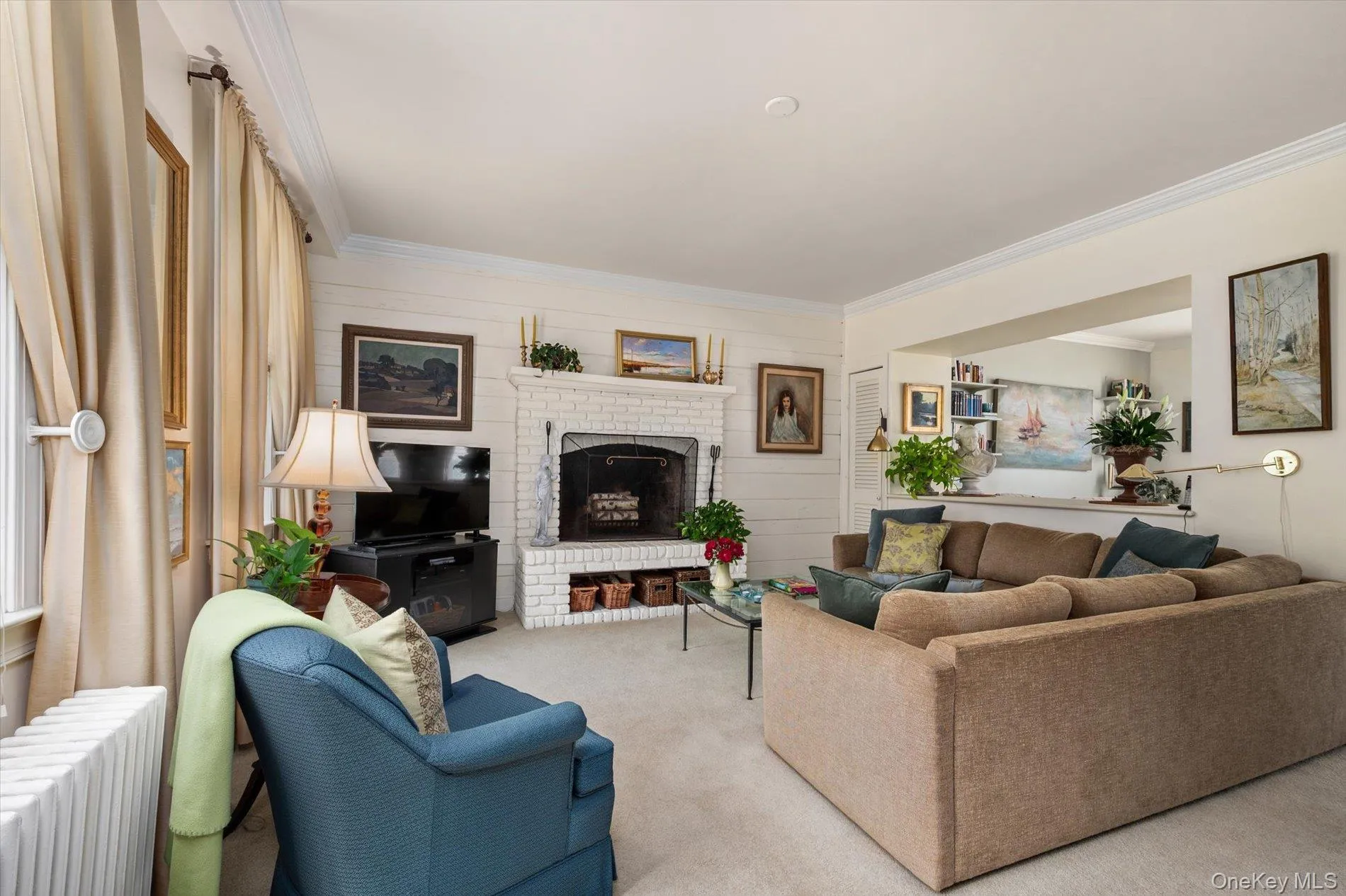 Living area featuring a white brick fireplace, shiplap accent wall, crown molding, and a pass-through opening to an additional room Living area featuring a white brick fireplace, shiplap accent wall, crown molding, and a pass-through opening to an additional room