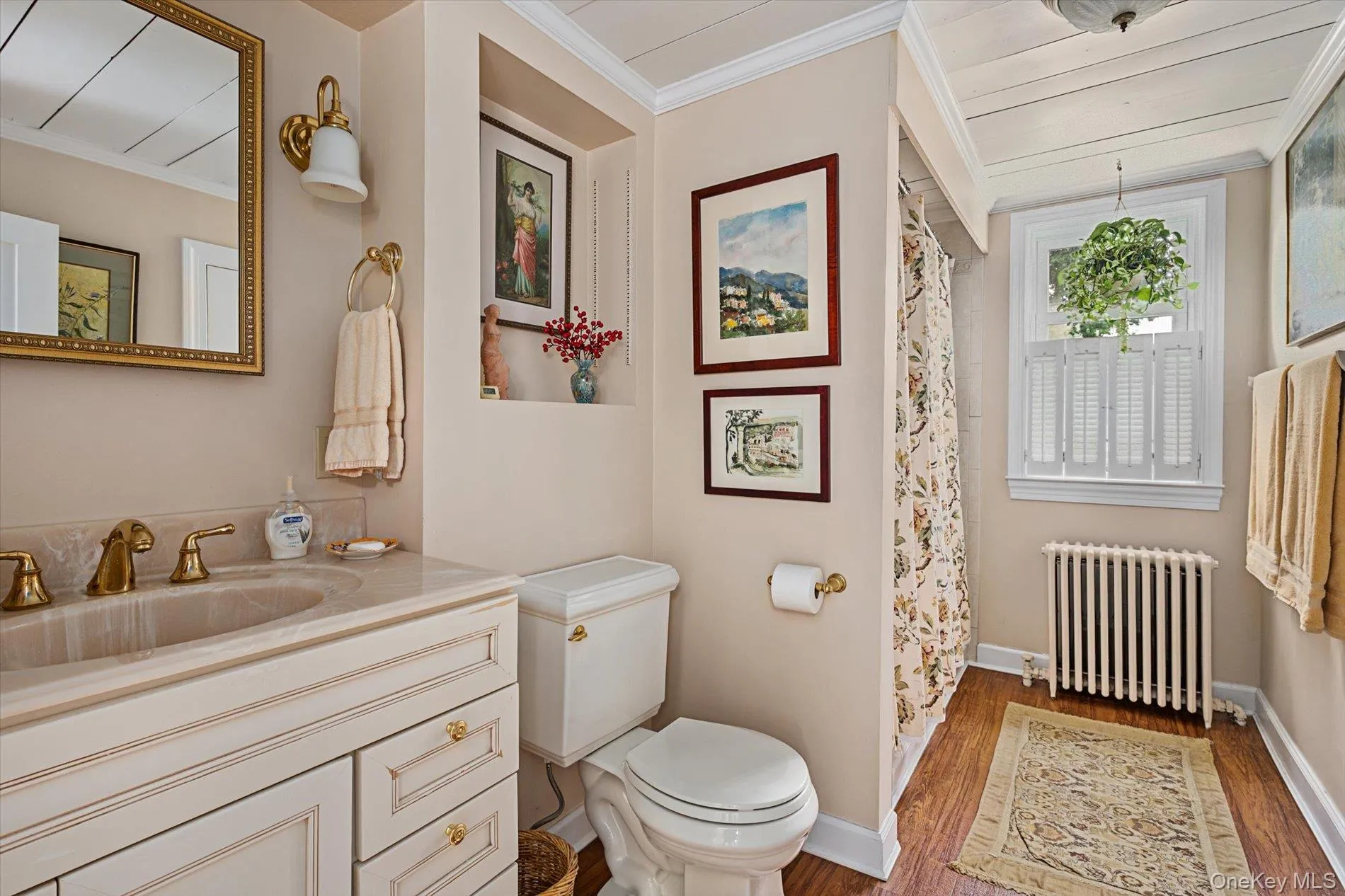 Bathroom featuring a vanity with a light-toned countertop, gold-toned fixtures, wood-finish flooring, and a window with plantation shutters Bathroom featuring a vanity with a light-toned countertop, gold-toned fixtures, wood-finish flooring, and a window with plantation shutters