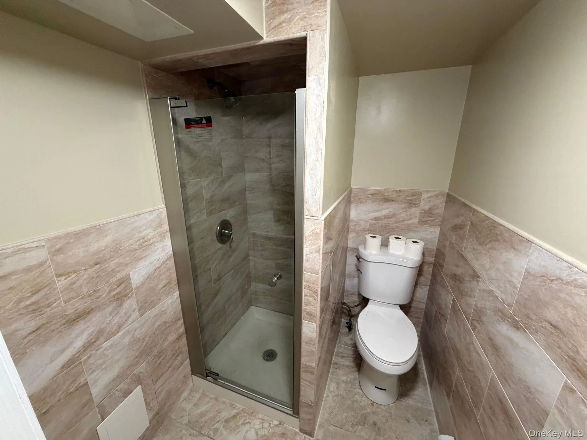 Full bathroom featuring a walk-in shower with a glass enclosure, a white commode, and extensive tile flooring and wainscoting Full bathroom featuring a walk-in shower with a glass enclosure, a white commode, and extensive tile flooring and wainscoting