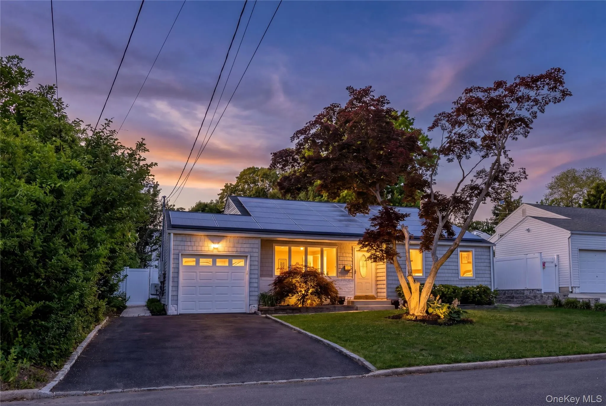 Shingle-sided residence featuring a paved driveway, mature landscaping, and a one-car garage with panel detailing Shingle-sided residence featuring a paved driveway, mature landscaping, and a one-car garage with panel detailing
