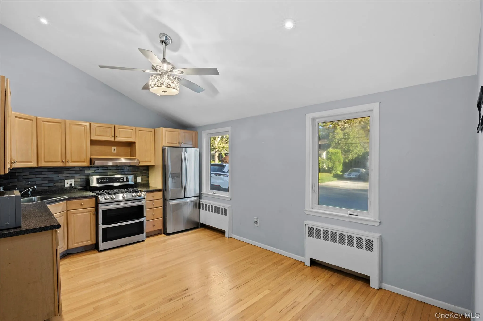 Kitchen featuring light-tone wood cabinetry, stainless steel appliances, dark countertop, and a dark tile backsplash Kitchen featuring light-tone wood cabinetry, stainless steel appliances, dark countertop, and a dark tile backsplash