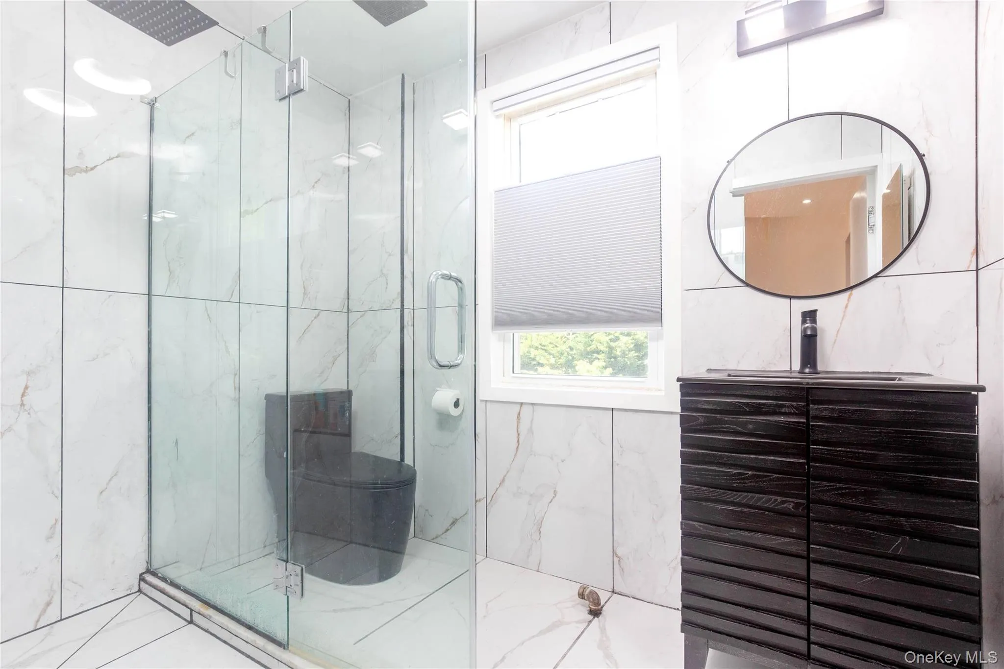 Modern bathroom featuring a glass-enclosed shower with a rainfall showerhead, white tiled walls with subtle veining, a dark wood-finish vanity with a vessel sink, and an oval mirror with a black frame Modern bathroom featuring a glass-enclosed shower with a rainfall showerhead, white tiled walls with subtle veining, a dark wood-finish vanity with a vessel sink, and an oval mirror with a black frame