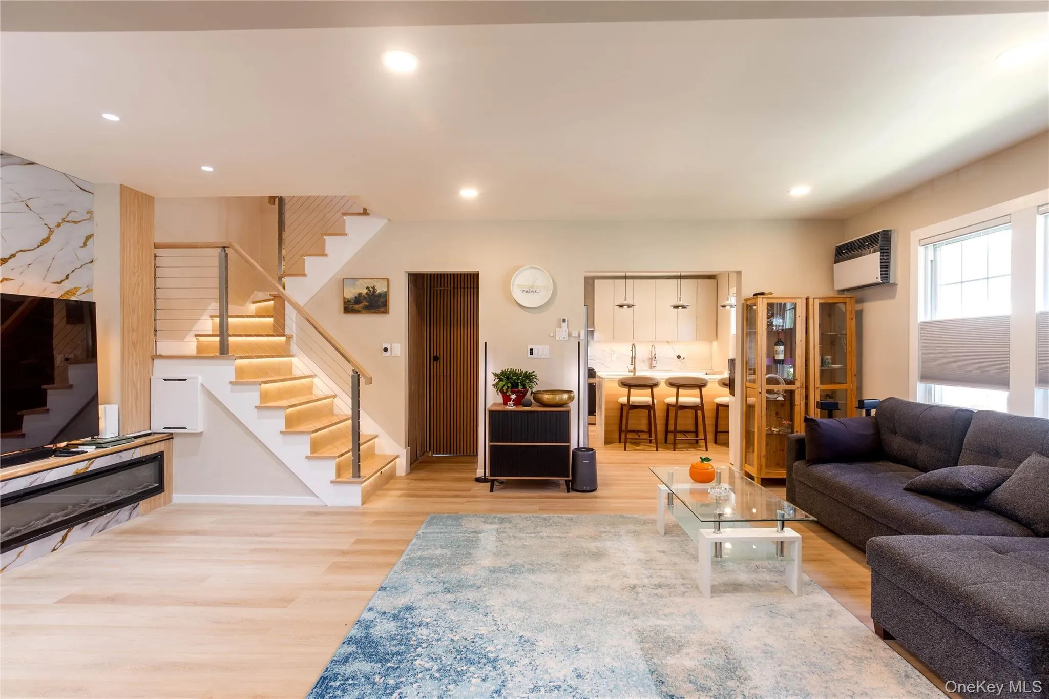 Open-concept living area featuring light wood-finish flooring, recessed ceiling lighting, a built-in linear fireplace with a marble-finish surround, and a modern staircase with cable railing Open-concept living area featuring light wood-finish flooring, recessed ceiling lighting, a built-in linear fireplace with a marble-finish surround, and a modern staircase with cable railing