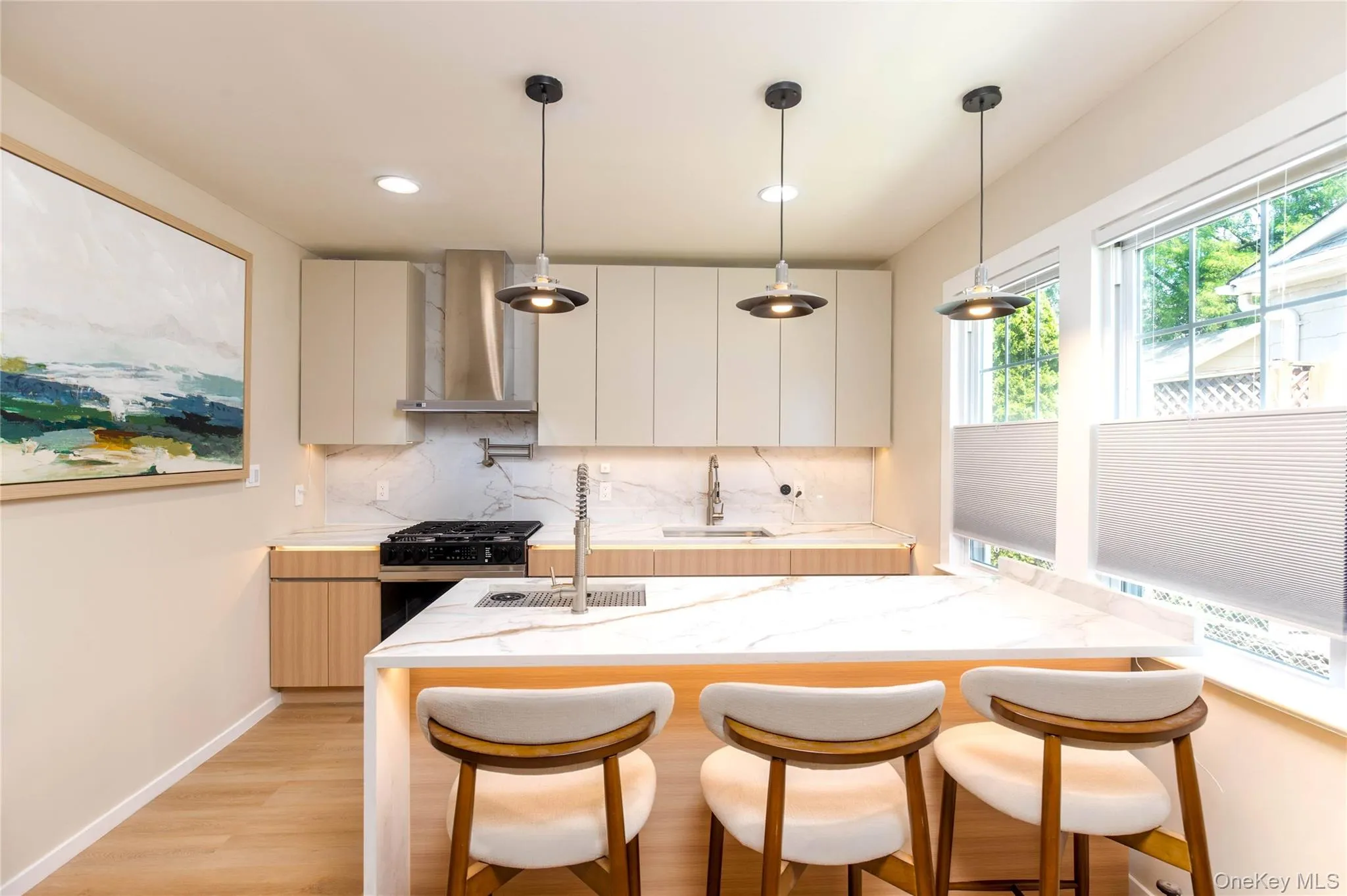 Modern kitchen featuring a waterfall island with integrated lighting, a gas range with a stainless steel range hood, and light-toned cabinetry Modern kitchen featuring a waterfall island with integrated lighting, a gas range with a stainless steel range hood, and light-toned cabinetry