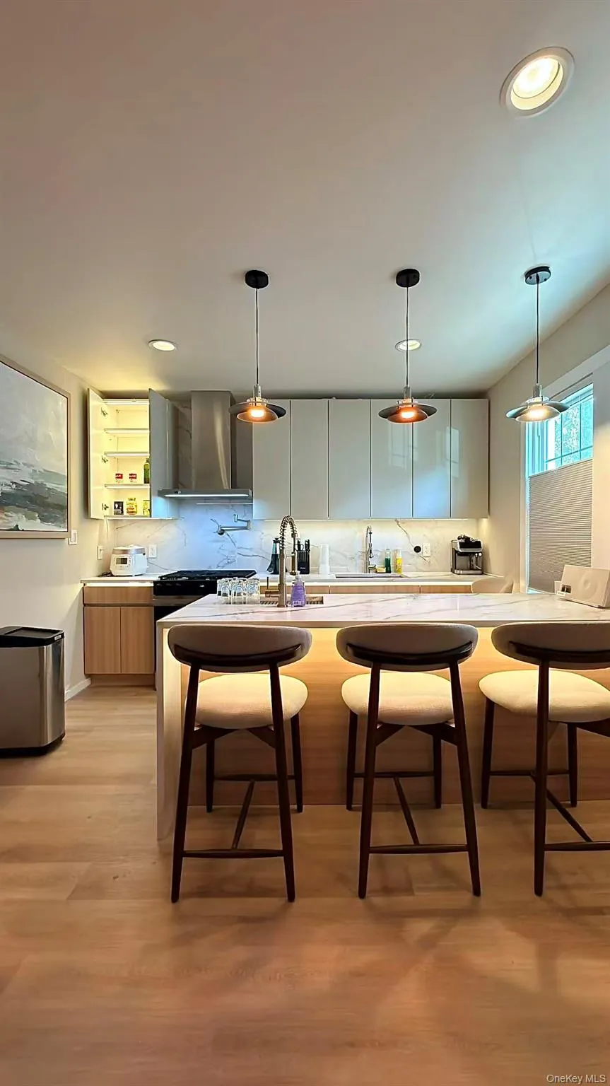 Modern kitchen featuring wood-finish flooring, a central island with integrated lighting, a stainless steel range hood, light-toned flat-panel cabinetry, and recessed lighting Modern kitchen featuring wood-finish flooring, a central island with integrated lighting, a stainless steel range hood, light-toned flat-panel cabinetry, and recessed lighting