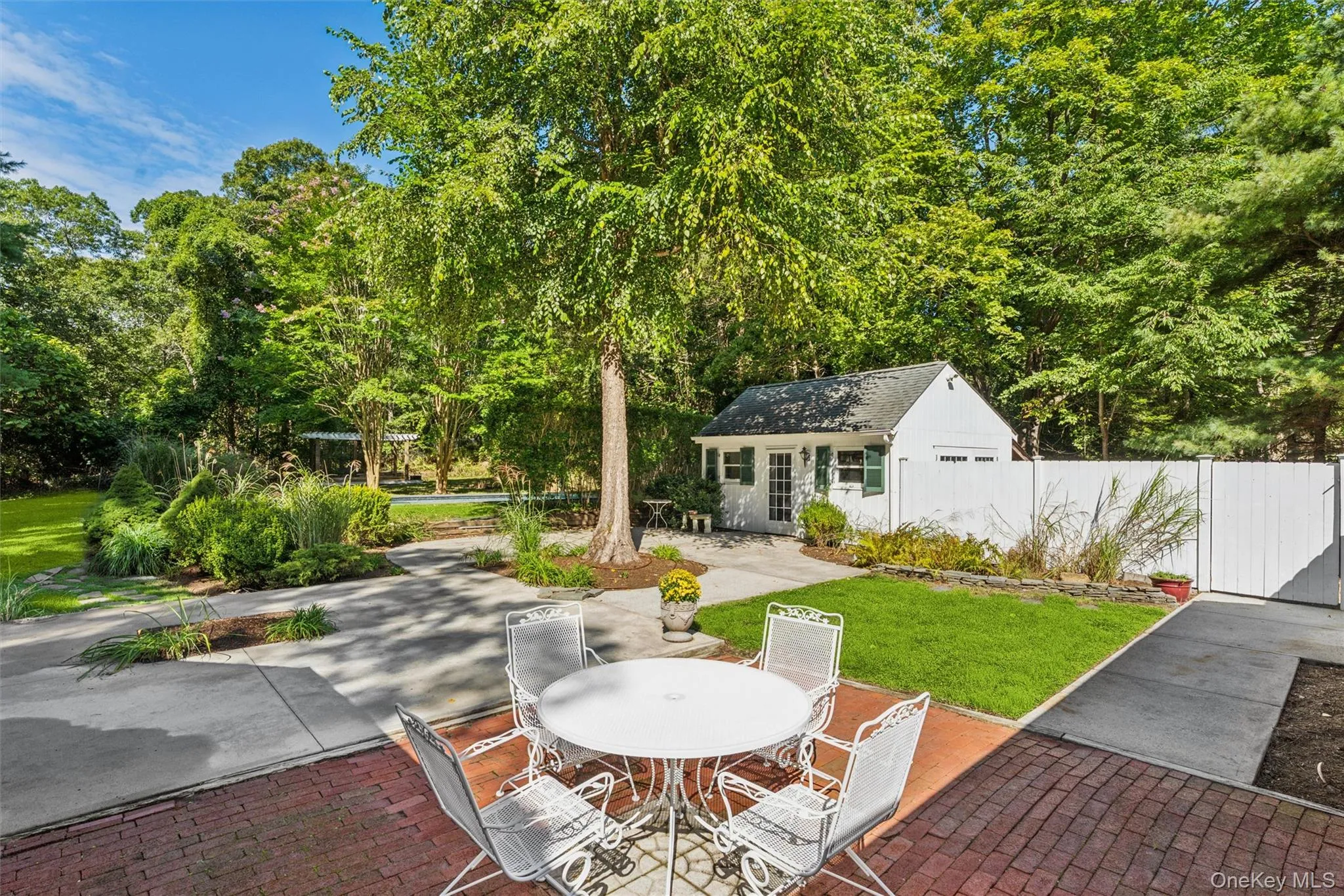Expansive backyard featuring a brick patio, concrete pathways, a detached outbuilding with a garage door and white entry door, and a white privacy fence Expansive backyard featuring a brick patio, concrete pathways, a detached outbuilding with a garage door and white entry door, and a white privacy fence