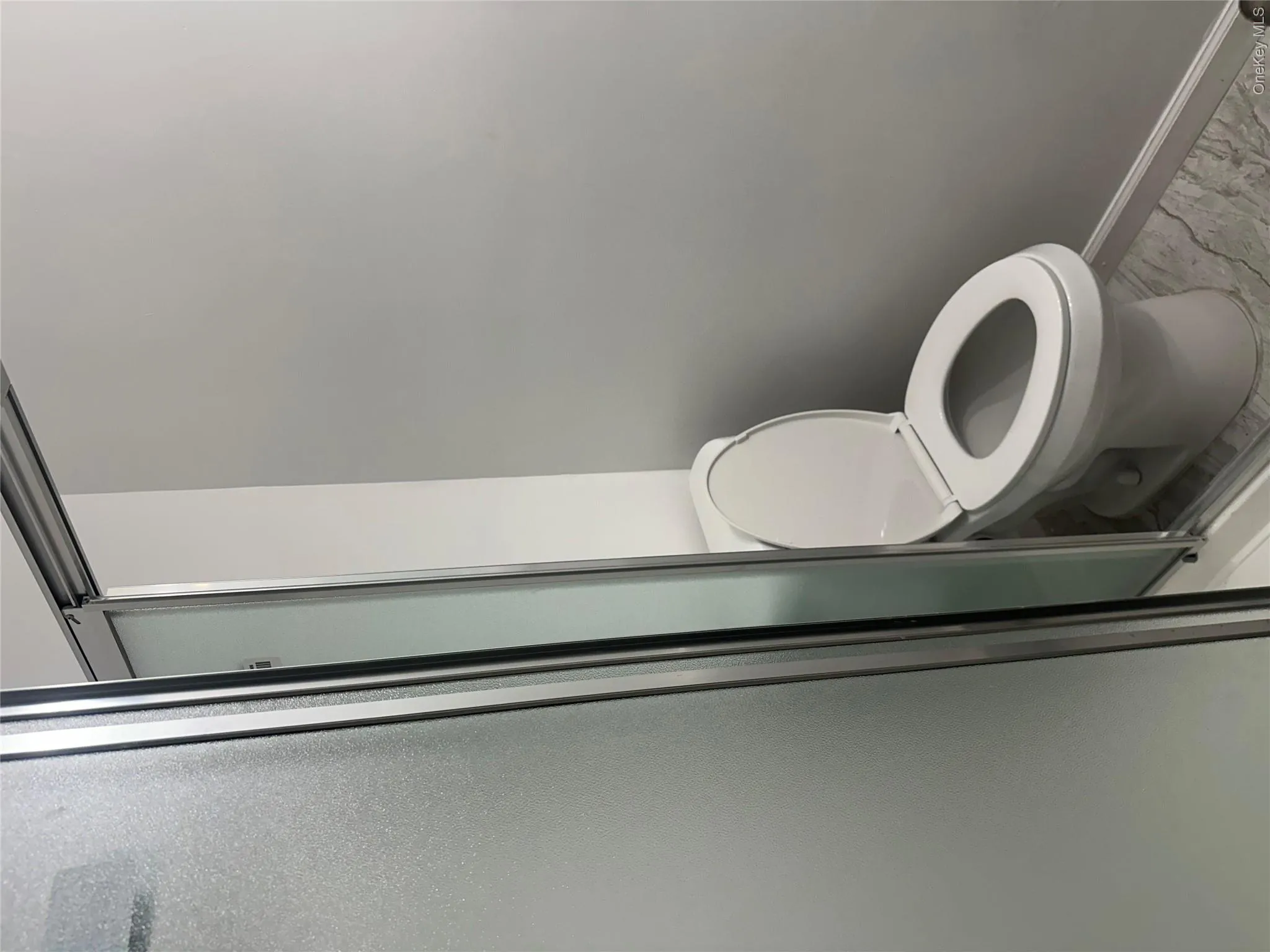 White commode with a round bowl set against light gray walls, featuring a frosted glass shower enclosure with chrome framing and patterned gray flooring White commode with a round bowl set against light gray walls, featuring a frosted glass shower enclosure with chrome framing and patterned gray flooring