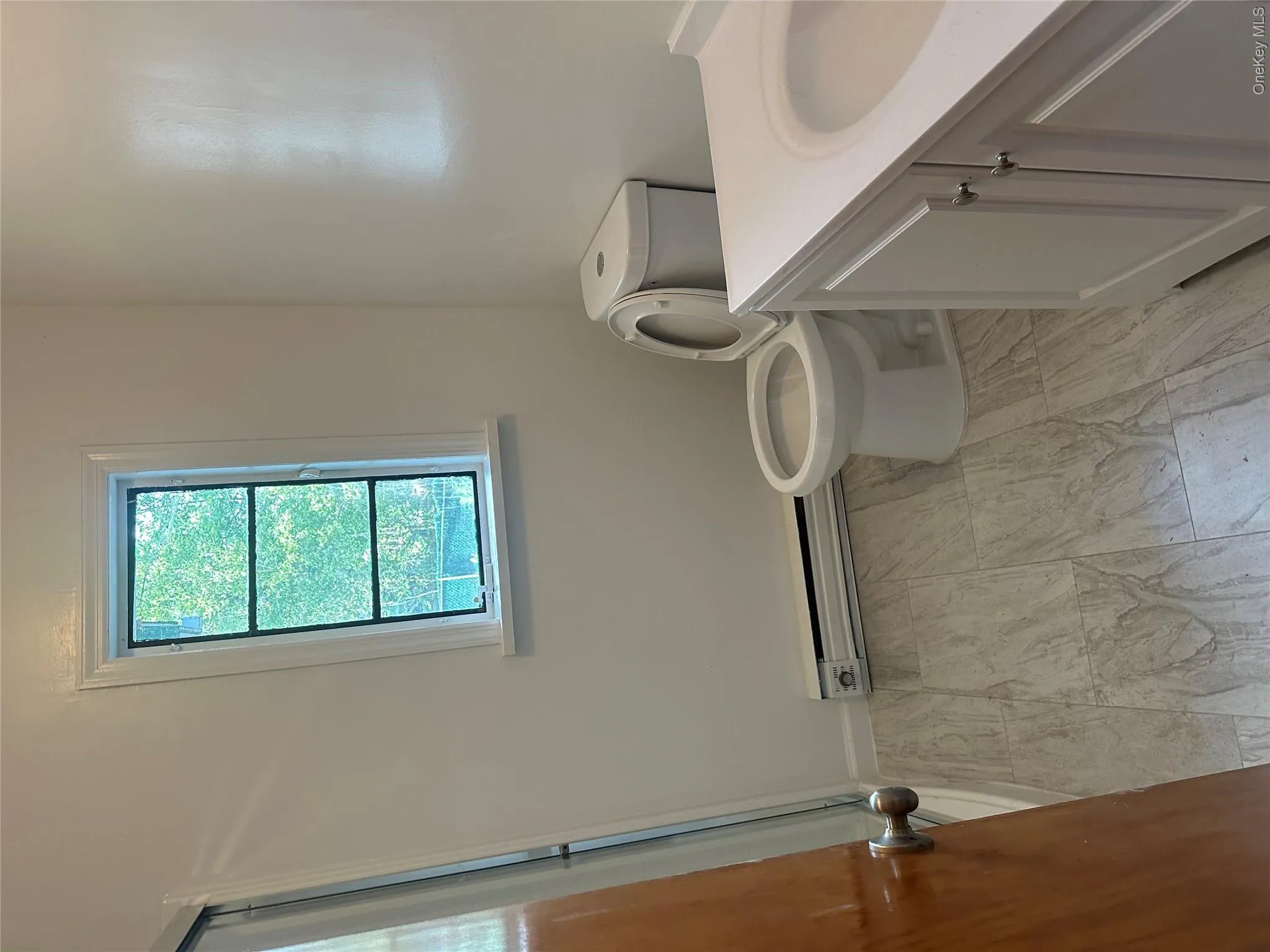 Bathroom featuring a white vanity with integrated sink, a commode, and light-toned, rectangular tile flooring Bathroom featuring a white vanity with integrated sink, a commode, and light-toned, rectangular tile flooring