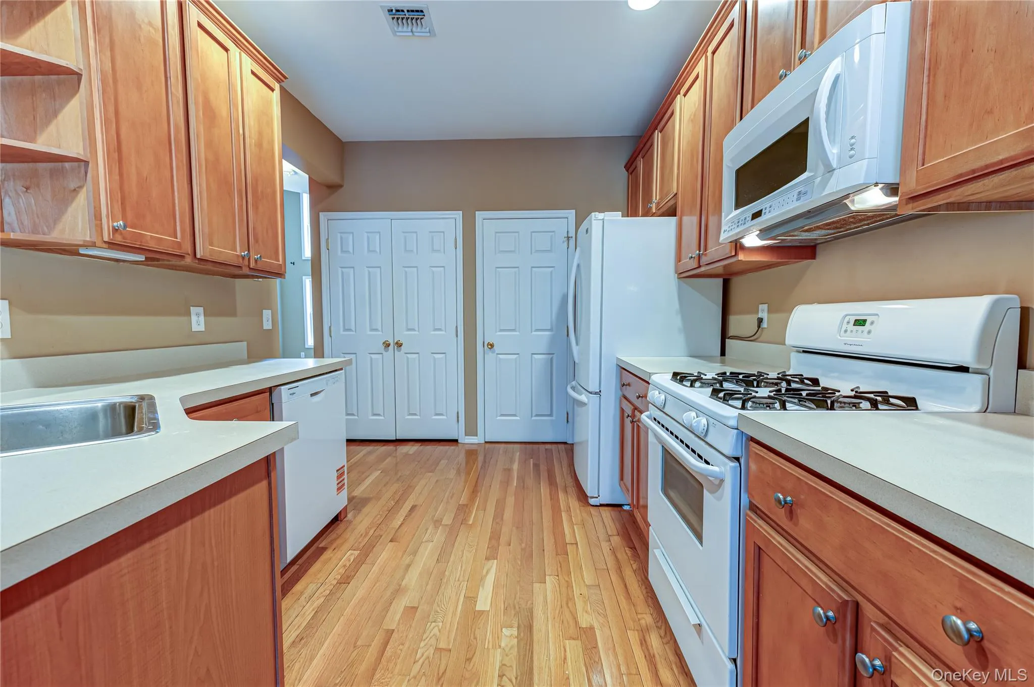 Galley kitchen featuring wood-finish cabinetry, light countertops, hardwood flooring, a stainless steel sink, and white appliances including a brand new white Whirlpool Dishwasher Galley kitchen featuring wood-finish cabinetry, light countertops, hardwood flooring, a stainless steel sink, and white appliances including a brand new white Whirlpool Dishwasher