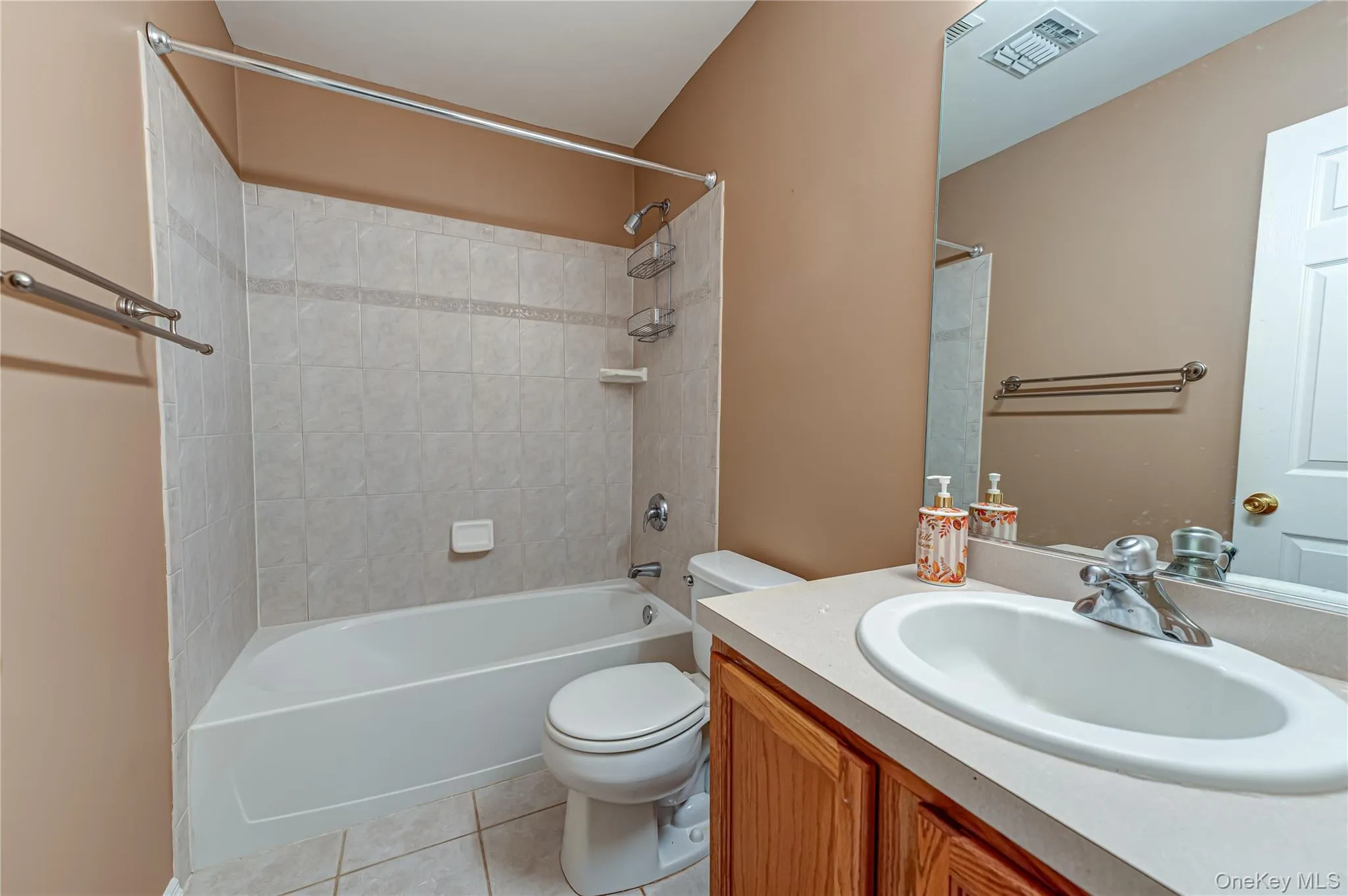 Full bathroom featuring a single vanity with wood-finish cabinetry and an integrated sink, a white bathtub with tile surround, and tile flooring Full bathroom featuring a single vanity with wood-finish cabinetry and an integrated sink, a white bathtub with tile surround, and tile flooring