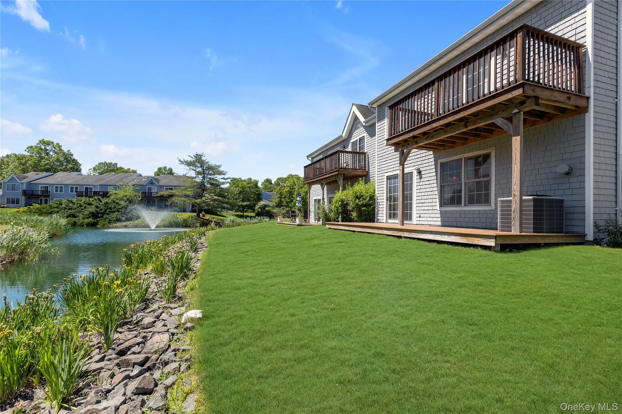 Shingle-sided exterior featuring a wooden balcony and ground-level deck, overlooking a manicured lawn and a body of water with a fountain Shingle-sided exterior featuring a wooden balcony and ground-level deck, overlooking a manicured lawn and a body of water with a fountain
