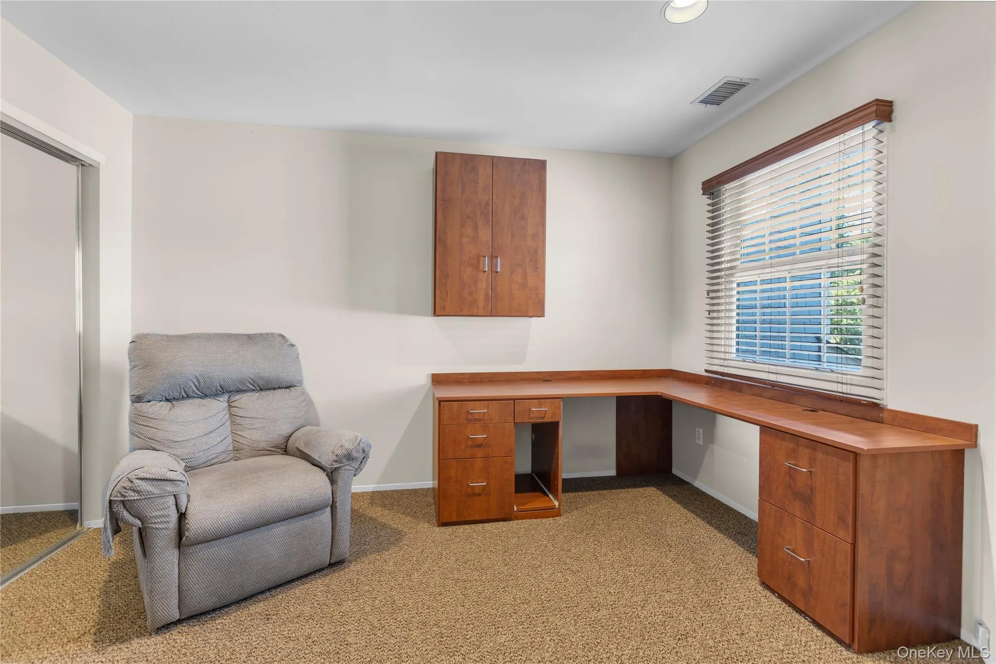 Room featuring a corner wood-finish desk with integrated cabinetry, a window with blinds, a wall-mounted wood-finish cabinet, and textured carpet flooring Room featuring a corner wood-finish desk with integrated cabinetry, a window with blinds, a wall-mounted wood-finish cabinet, and textured carpet flooring