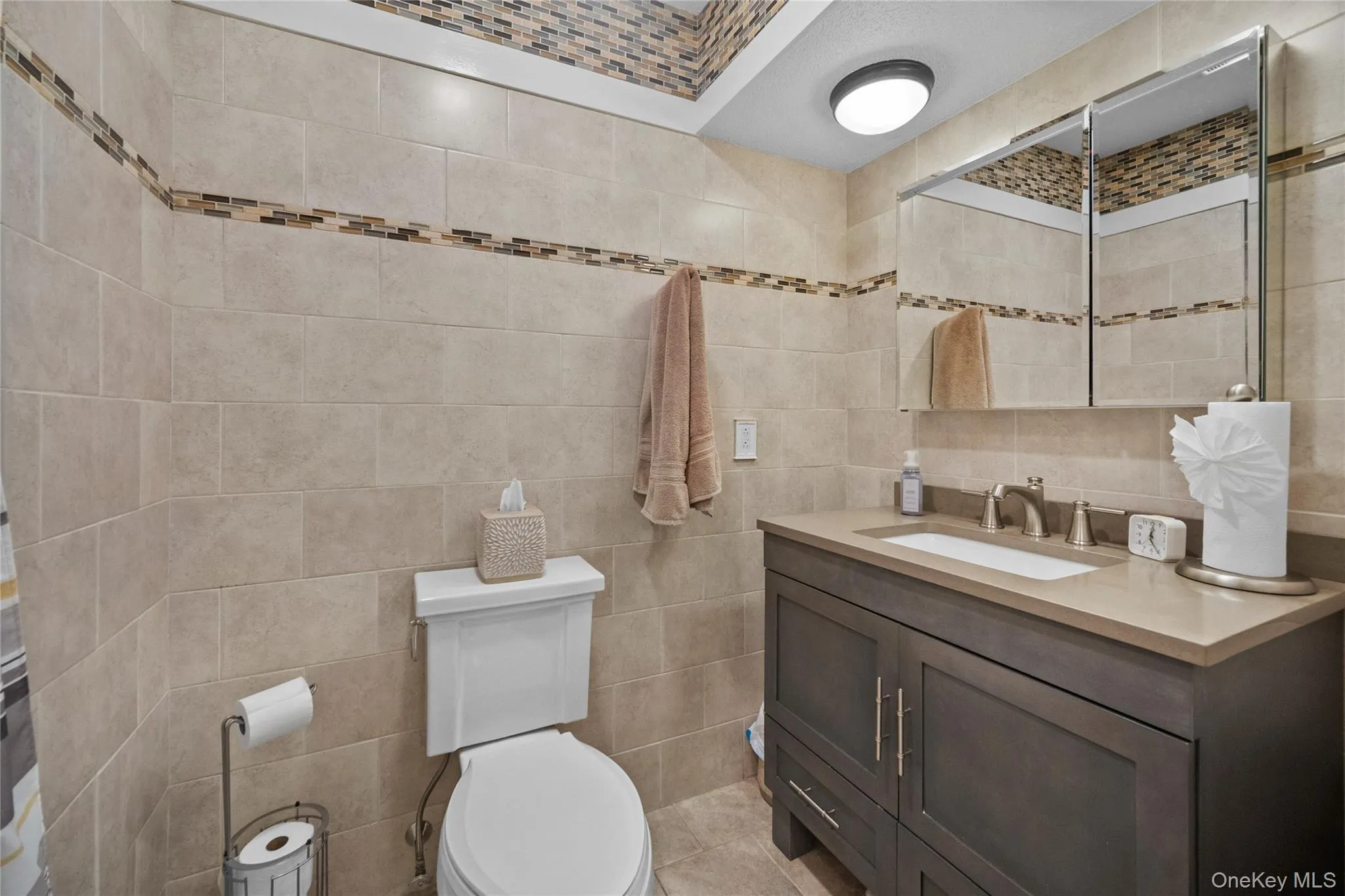 Bathroom featuring floor-to-ceiling tile, a decorative tile accent band, a white toilet, a wood-finish vanity with an integrated sink, and a mirrored medicine cabinet Bathroom featuring floor-to-ceiling tile, a decorative tile accent band, a white toilet, a wood-finish vanity with an integrated sink, and a mirrored medicine cabinet