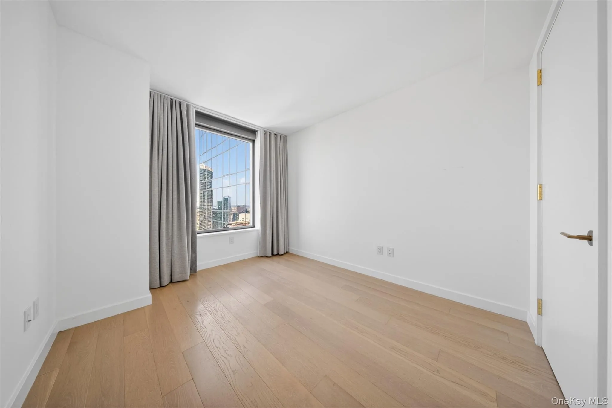 Room featuring wood-finish flooring, a full-height window with gray drapery, high ceilings, white walls, and a white interior door with brass hardware Room featuring wood-finish flooring, a full-height window with gray drapery, high ceilings, white walls, and a white interior door with brass hardware