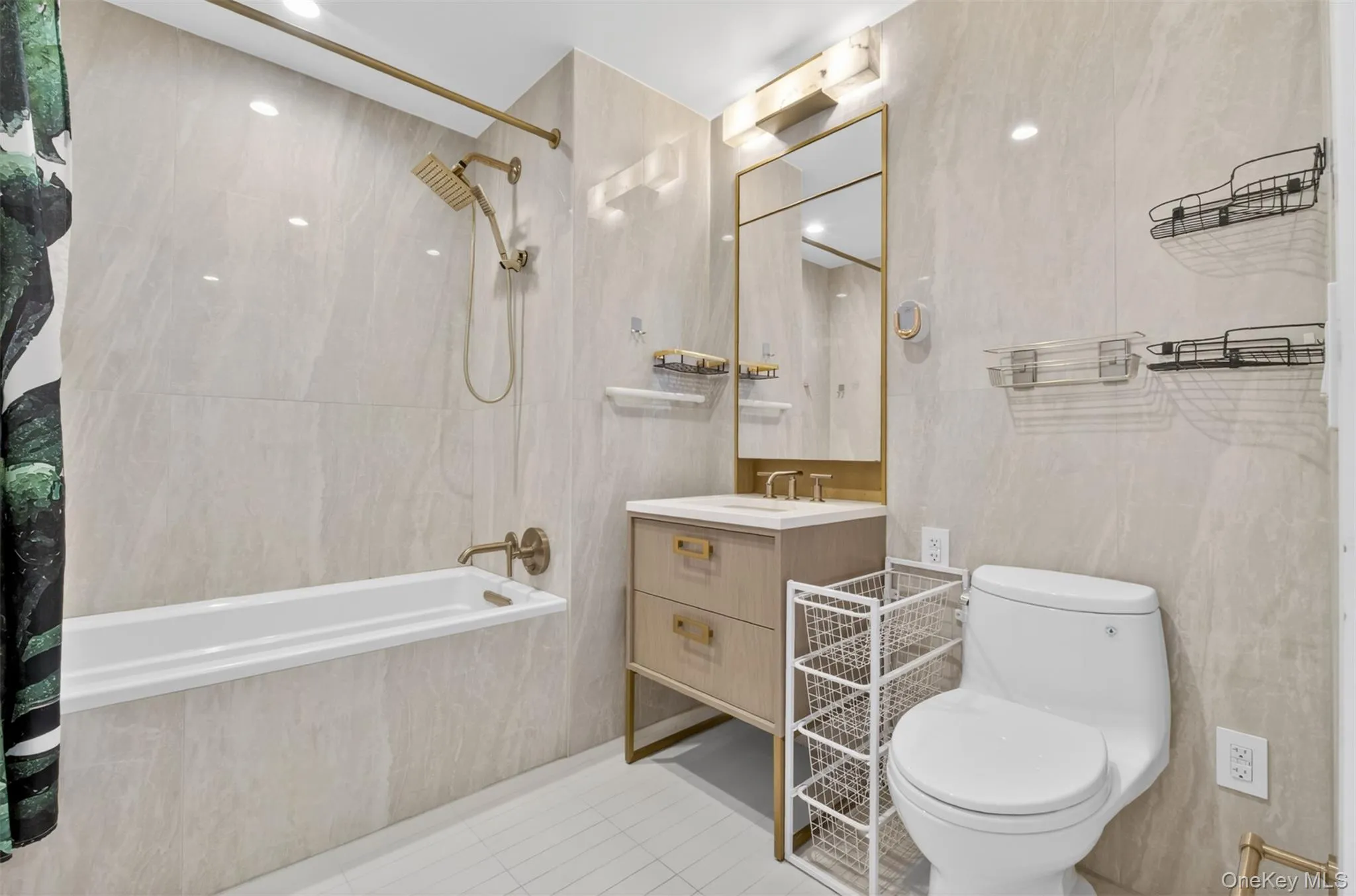 Bathroom featuring full-height light-toned tile, a white bathtub with gold-finish fixtures, a vanity with gold hardware, and a framed mirror with integrated lighting Bathroom featuring full-height light-toned tile, a white bathtub with gold-finish fixtures, a vanity with gold hardware, and a framed mirror with integrated lighting