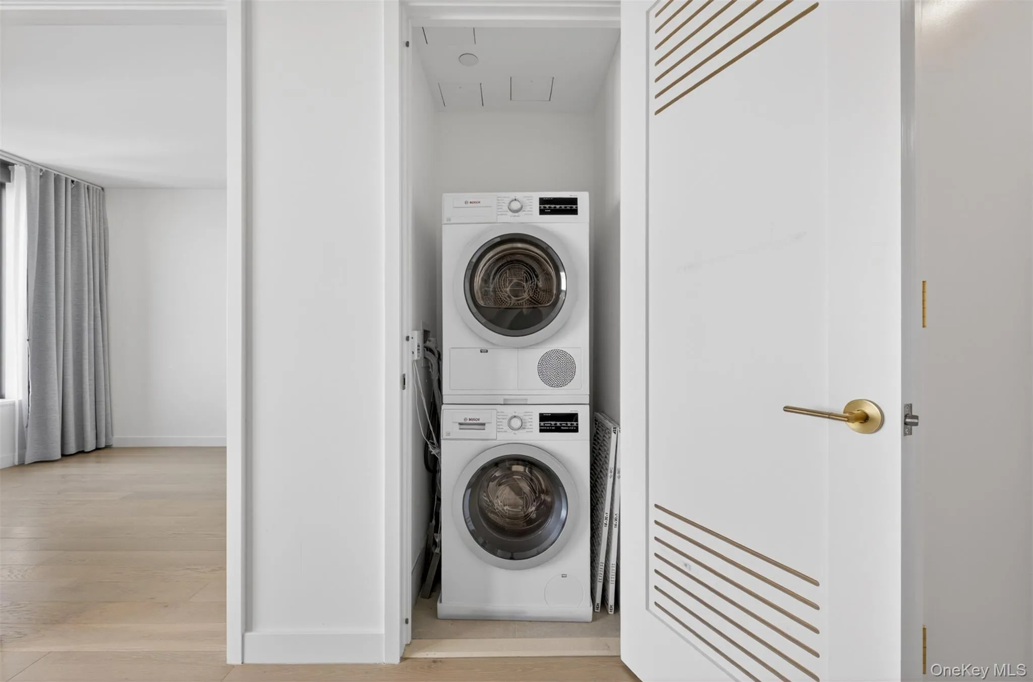 Laundry closet with stacked washer and dryer appliances, featuring a white door with decorative gold-toned detailing and a gold-toned lever handle Laundry closet with stacked washer and dryer appliances, featuring a white door with decorative gold-toned detailing and a gold-toned lever handle