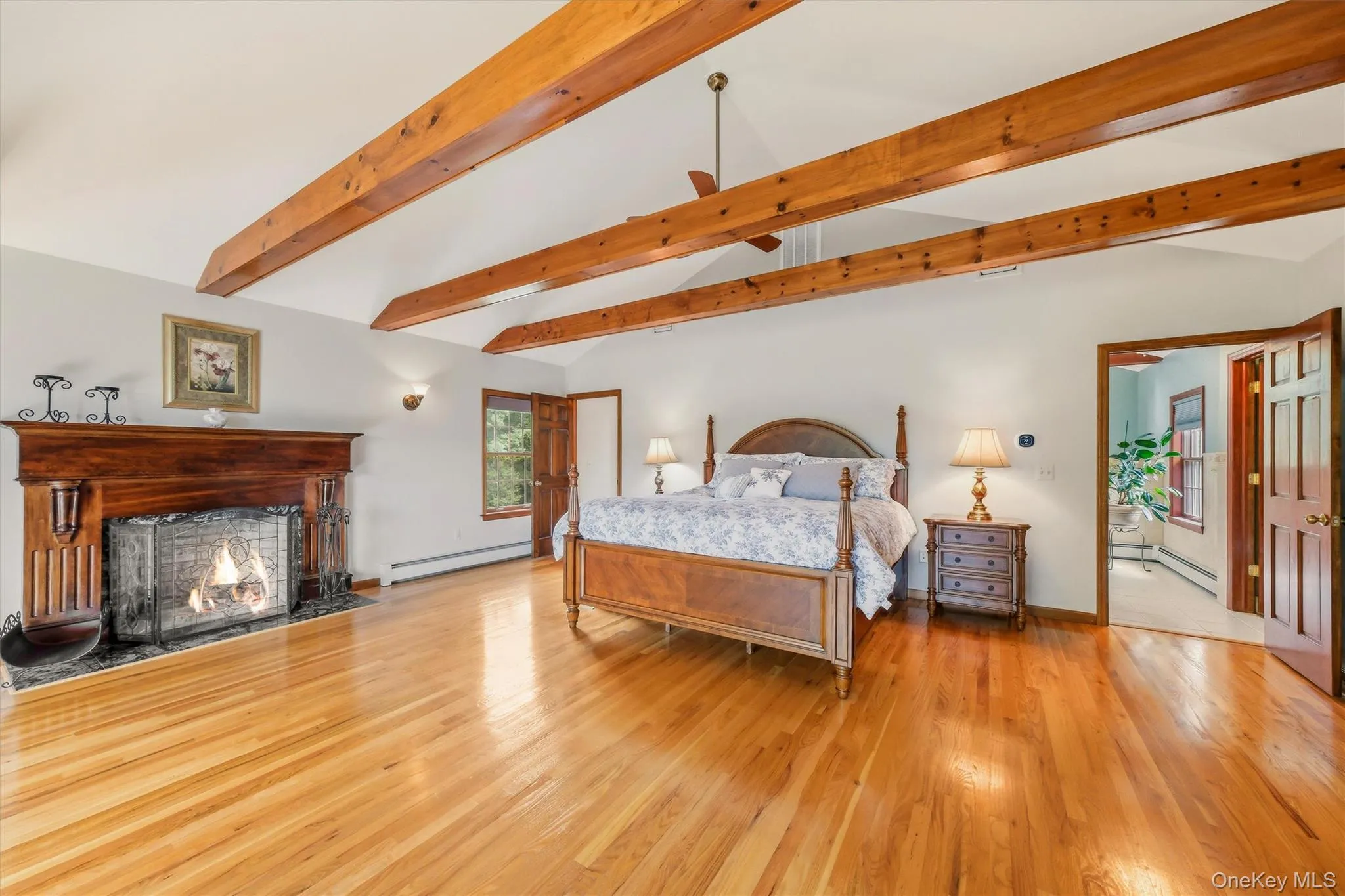 Spacious room featuring a vaulted ceiling with exposed wood beams, a prominent wood-mantled fireplace with a stone surround, polished wood-finish flooring, and a ceiling fan Spacious room featuring a vaulted ceiling with exposed wood beams, a prominent wood-mantled fireplace with a stone surround, polished wood-finish flooring, and a ceiling fan
