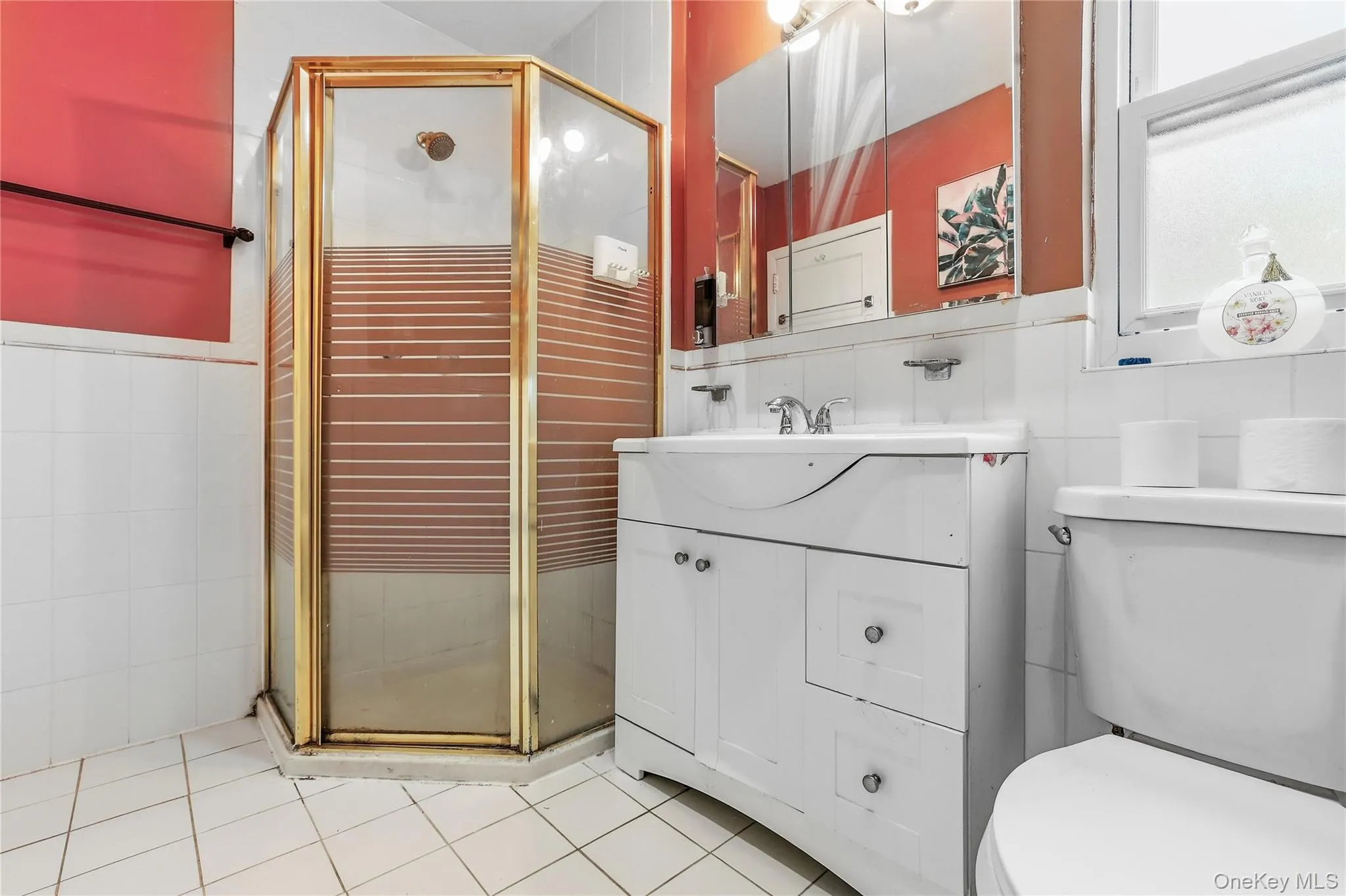 Bathroom featuring an enclosed corner shower with a gold-finish frame, a white vanity with integrated sink, a mirrored medicine cabinet, a window, and white square floor tiling Bathroom featuring an enclosed corner shower with a gold-finish frame, a white vanity with integrated sink, a mirrored medicine cabinet, a window, and white square floor tiling