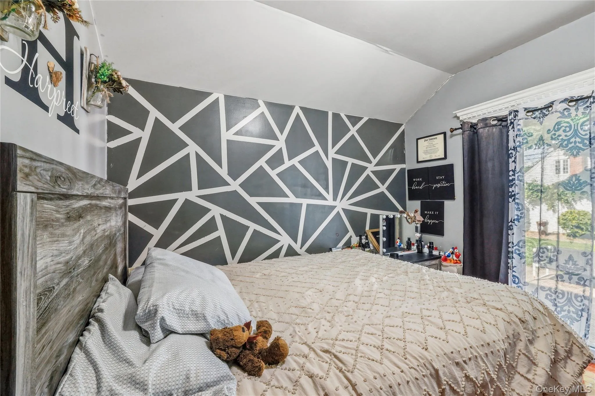 Bedroom featuring a wood-finish headboard, a geometric accent wall, and a window with decorative curtains Bedroom featuring a wood-finish headboard, a geometric accent wall, and a window with decorative curtains