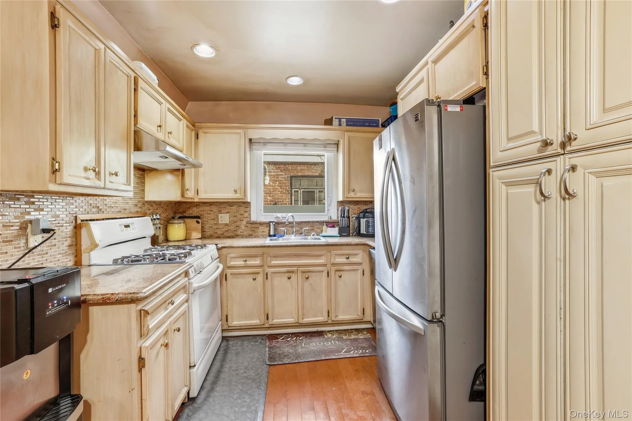 Kitchen featuring light wood cabinetry, a mosaic tile backsplash, a stainless steel refrigerator, and built-in overhead lighting Kitchen featuring light wood cabinetry, a mosaic tile backsplash, a stainless steel refrigerator, and built-in overhead lighting