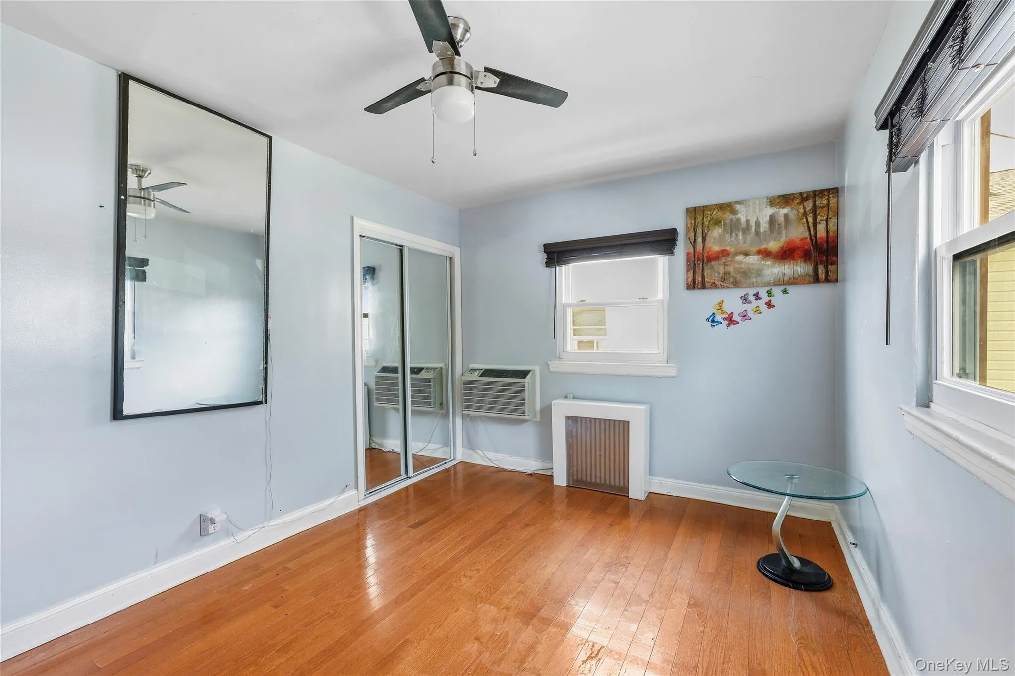 Room featuring wood-finish flooring, a ceiling fan, sliding mirror closet doors, and multiple windows with blinds Room featuring wood-finish flooring, a ceiling fan, sliding mirror closet doors, and multiple windows with blinds