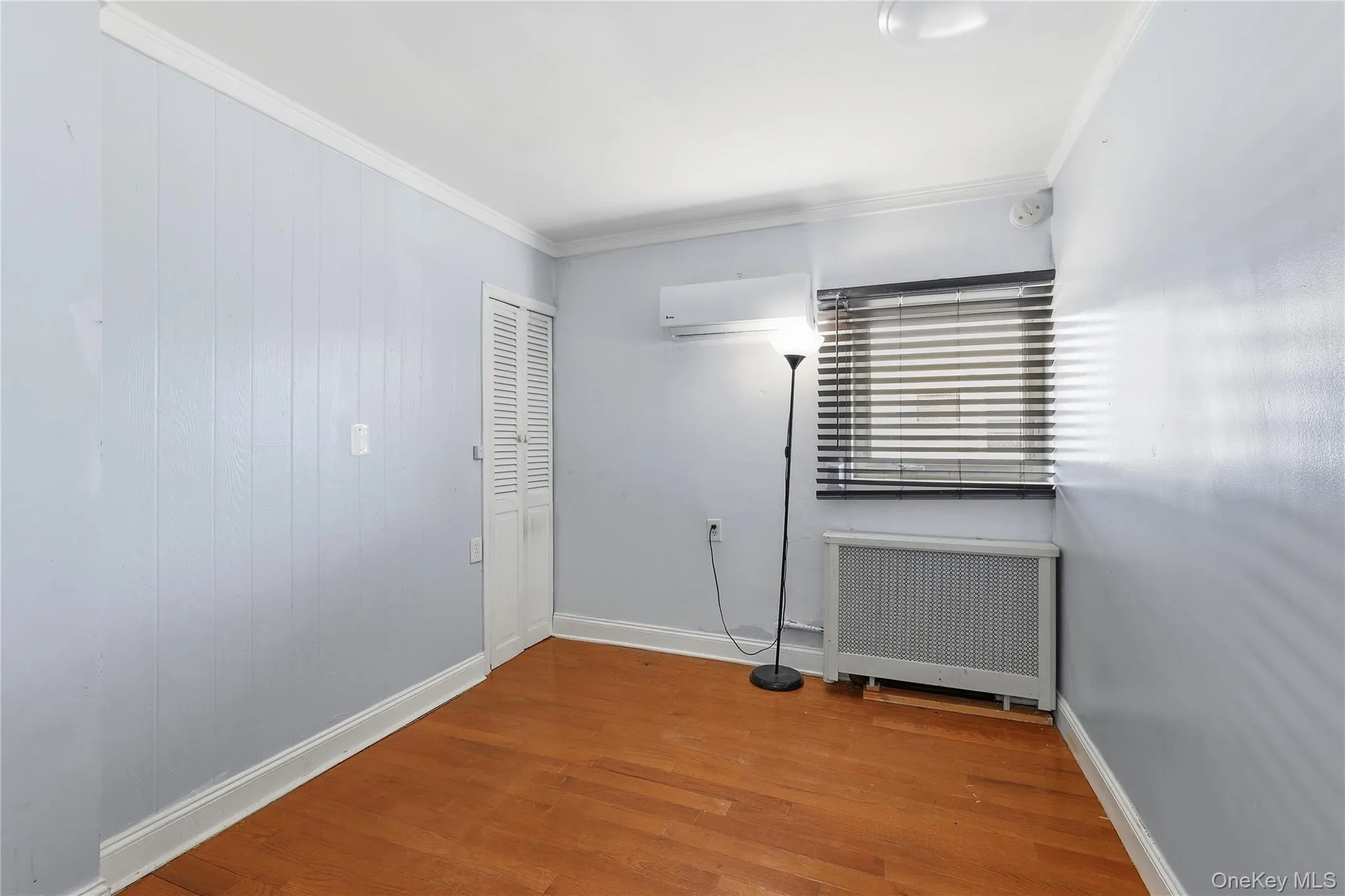 Second floor Bedroom 2 featuring wood-finish flooring, light blue paneled walls, white trim, a louvered bi-fold door, and a window with horizontal blinds Second floor Bedroom 2 featuring wood-finish flooring, light blue paneled walls, white trim, a louvered bi-fold door, and a window with horizontal blinds