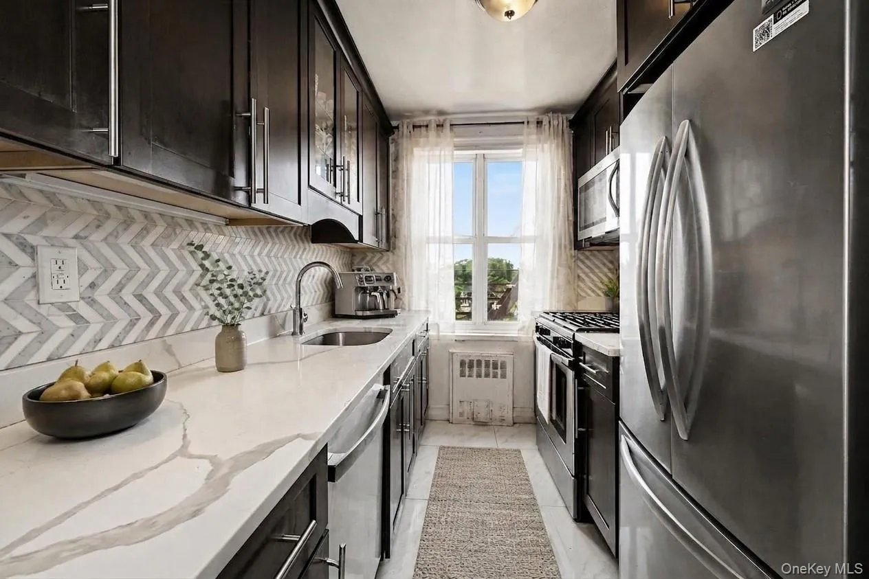 Galley kitchen featuring dark wood cabinetry, stainless steel appliances, white countertops, and a chevron pattern backsplash. Virtually Enhanced. Galley kitchen featuring dark wood cabinetry, stainless steel appliances, white countertops, and a chevron pattern backsplash. Virtually Enhanced.