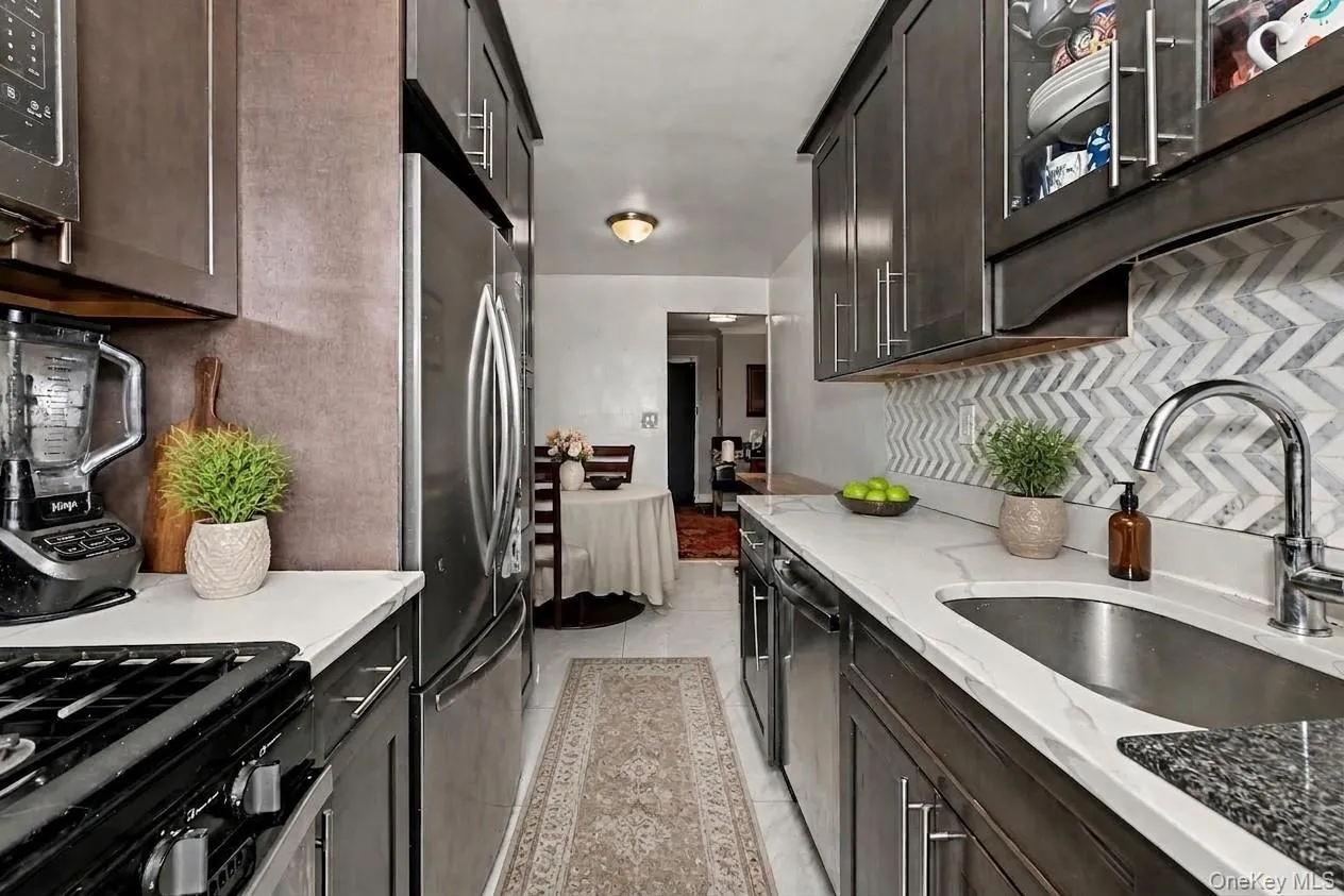 Galley kitchen featuring dark wood cabinetry, stainless steel appliances, white countertops, and a chevron pattern backsplash. Virtually Enhanced. Galley kitchen featuring dark wood cabinetry, stainless steel appliances, white countertops, and a chevron pattern backsplash. Virtually Enhanced.