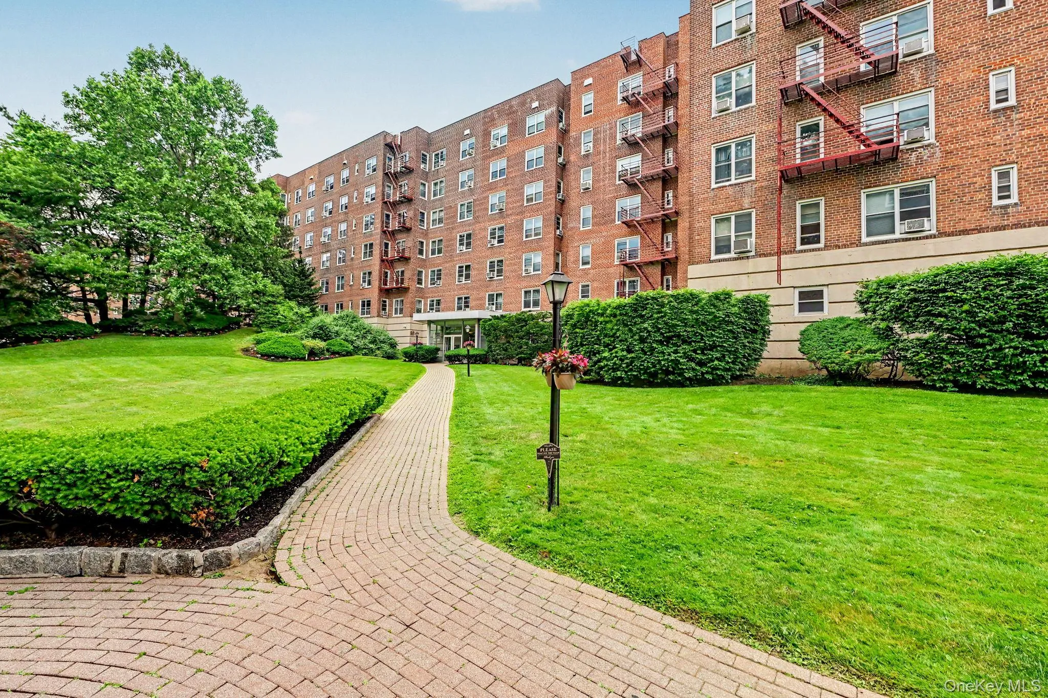 Brick multi-story building featuring multiple windows, exterior fire escapes, a recessed main entrance, and surrounding green landscaping Brick multi-story building featuring multiple windows, exterior fire escapes, a recessed main entrance, and surrounding green landscaping