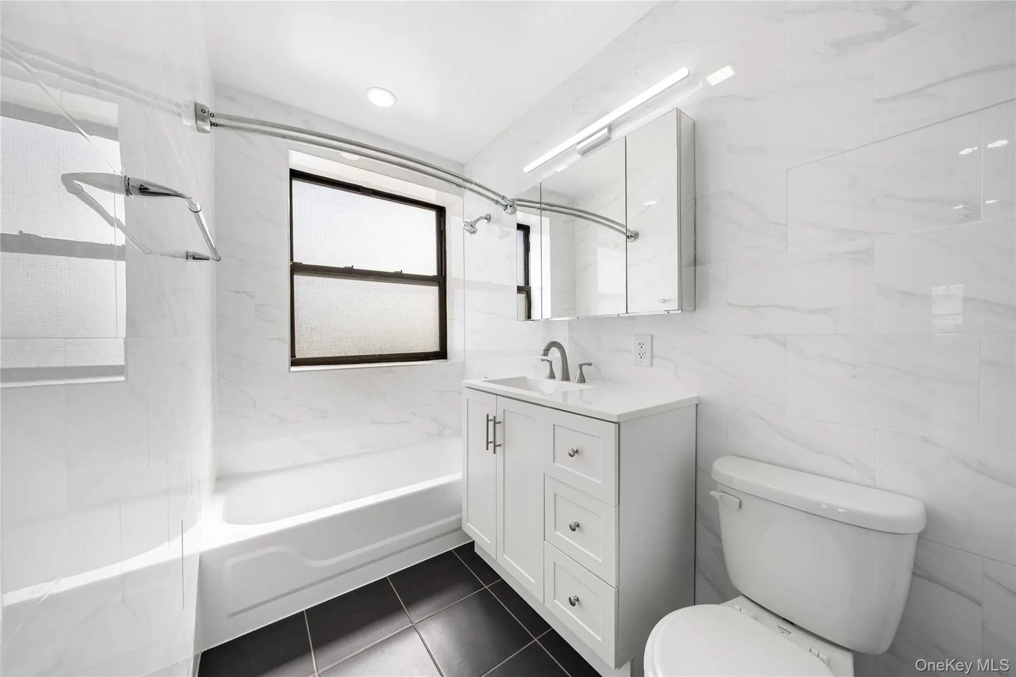 Bathroom featuring white subway tile walls, a white vanity with brushed nickel hardware, a white bathtub, a recessed medicine cabinet with integrated lighting, and dark tile flooring Bathroom featuring white subway tile walls, a white vanity with brushed nickel hardware, a white bathtub, a recessed medicine cabinet with integrated lighting, and dark tile flooring