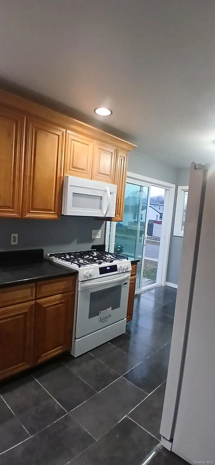 Kitchen with white appliances, dark countertops, brown cabinets, recessed lighting, and dark tile patterned floors Kitchen with white appliances, dark countertops, brown cabinets, recessed lighting, and dark tile patterned floors