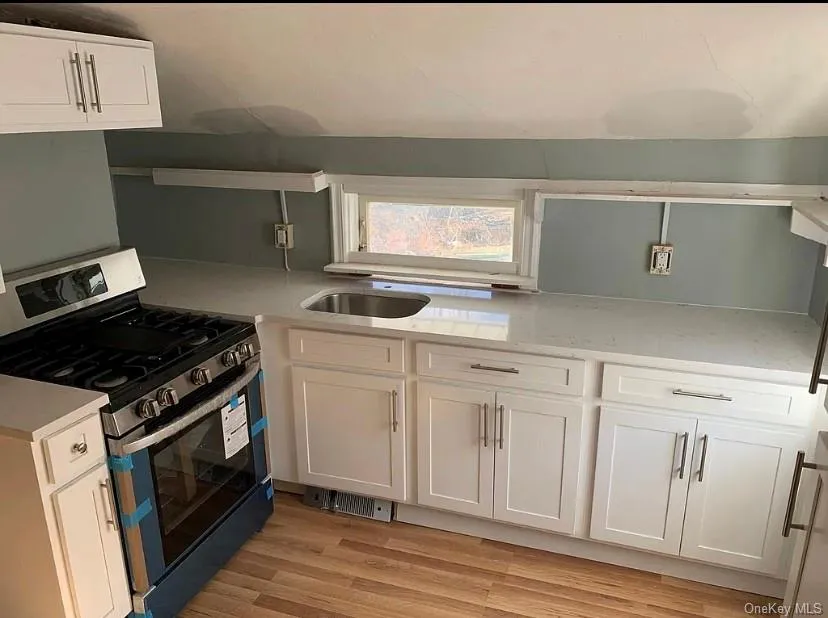 Compact kitchen featuring white shaker-style cabinetry, a stainless steel gas range, a stainless steel sink, light-toned countertops, and wood-finish flooring Compact kitchen featuring white shaker-style cabinetry, a stainless steel gas range, a stainless steel sink, light-toned countertops, and wood-finish flooring