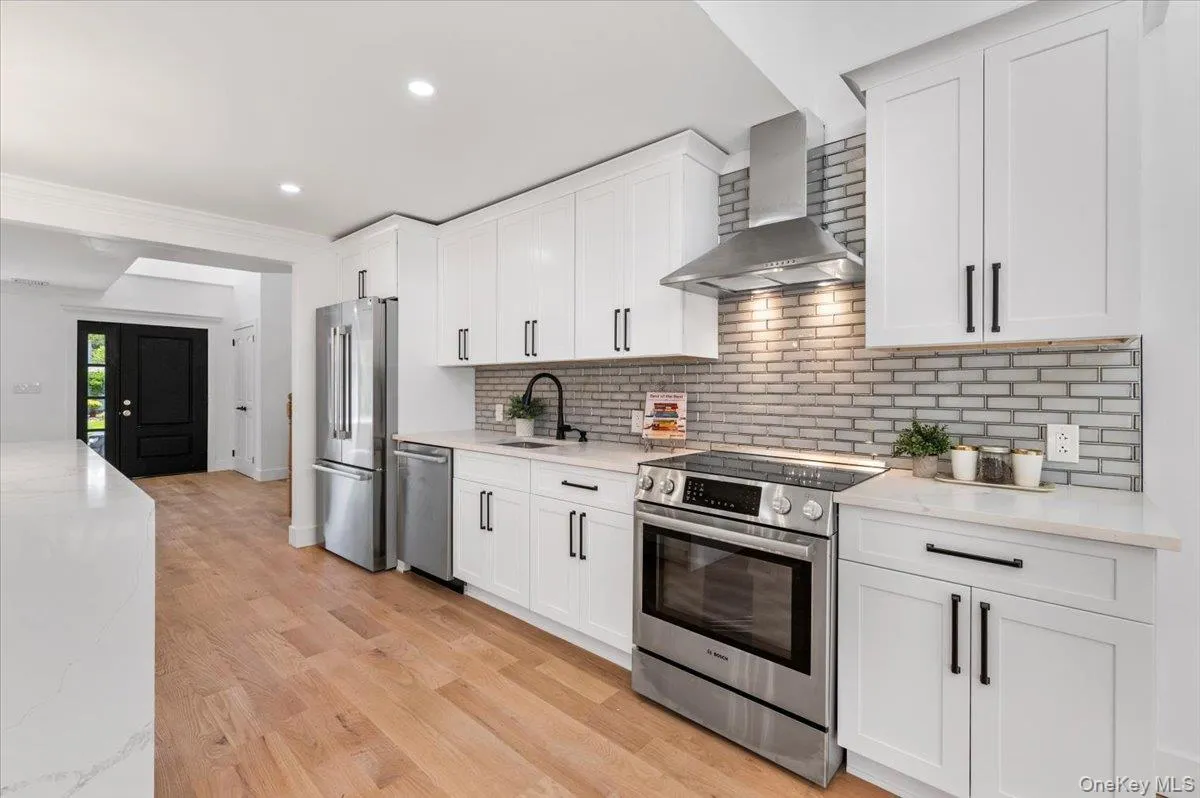 Kitchen with wall chimney range hood, stainless steel appliances, white cabinetry, backsplash, and recessed lighting Kitchen with wall chimney range hood, stainless steel appliances, white cabinetry, backsplash, and recessed lighting