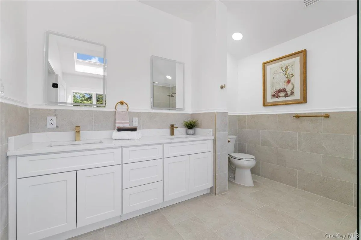 Full bathroom featuring tile walls, double vanity, tile patterned floors, a wainscoted wall, and a skylight Full bathroom featuring tile walls, double vanity, tile patterned floors, a wainscoted wall, and a skylight