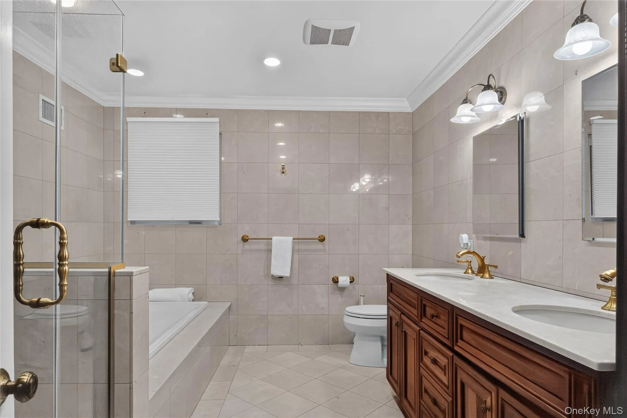 Massive hall bathroom featuring a dual-sink vanity with wood cabinetry and light-toned countertops, a built-in soaking tub, a glass-enclosed shower, and full-height wall tiling. Massive hall bathroom featuring a dual-sink vanity with wood cabinetry and light-toned countertops, a built-in soaking tub, a glass-enclosed shower, and full-height wall tiling.