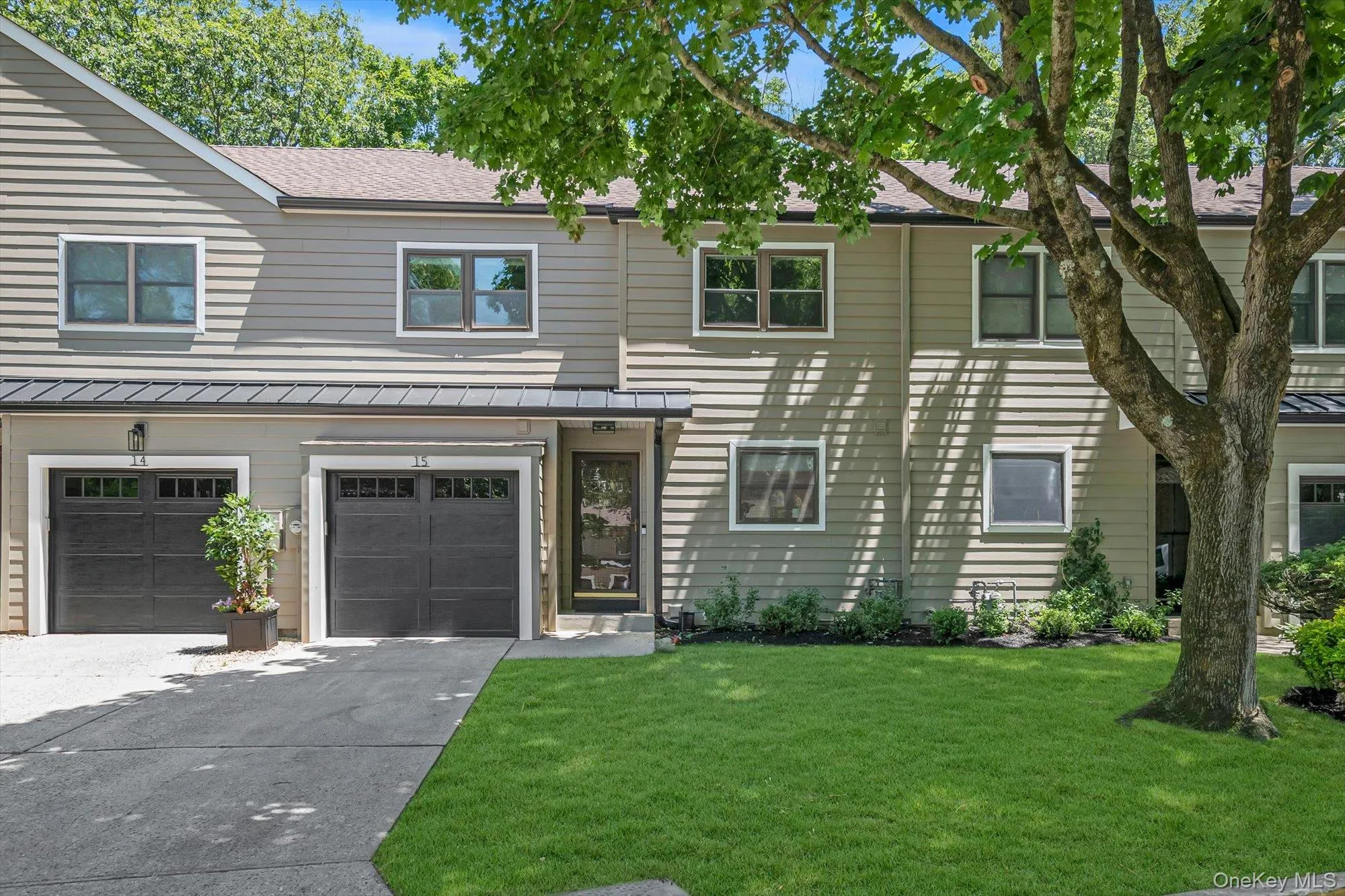Two-story townhome featuring neutral horizontal siding, dark garage doors with window inserts, a paved driveway, a manicured lawn, and mature trees Two-story townhome featuring neutral horizontal siding, dark garage doors with window inserts, a paved driveway, a manicured lawn, and mature trees