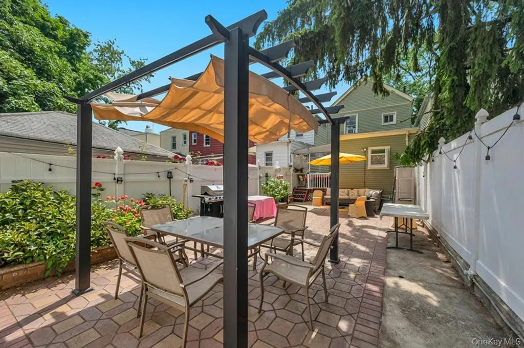 Paved patio area featuring a pergola with retractable canvas shades, a privacy fence, and mature trees providing natural shading Paved patio area featuring a pergola with retractable canvas shades, a privacy fence, and mature trees providing natural shading