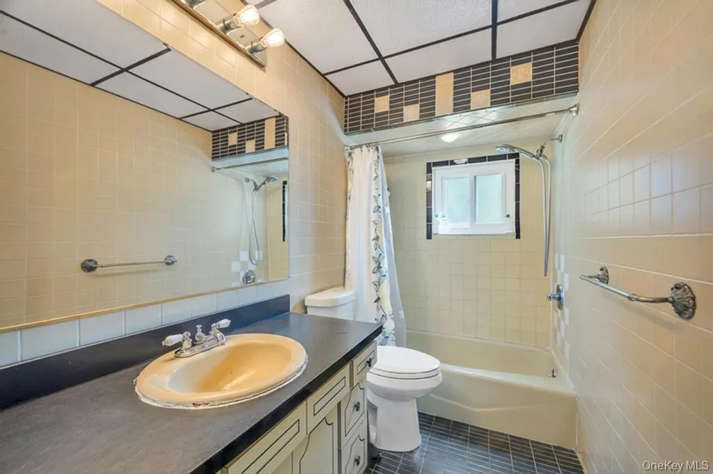 Full bathroom featuring a vanity with a surface-mounted sink, a large wall-mounted mirror, overhead lighting, tiled walls, and a shower-over-tub combination with a window Full bathroom featuring a vanity with a surface-mounted sink, a large wall-mounted mirror, overhead lighting, tiled walls, and a shower-over-tub combination with a window