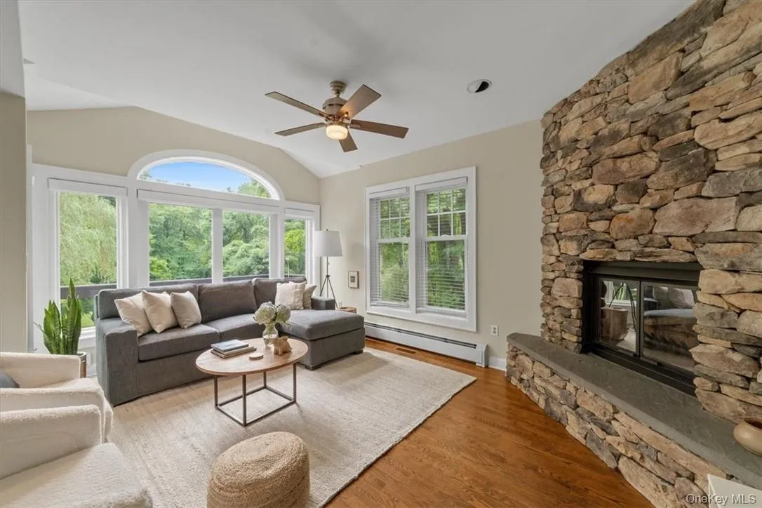 Living room with a fireplace, hardwood / wood-style flooring, ceiling fan, vaulted ceiling, and baseboard heating Living room with a fireplace, hardwood / wood-style flooring, ceiling fan, vaulted ceiling, and baseboard heating