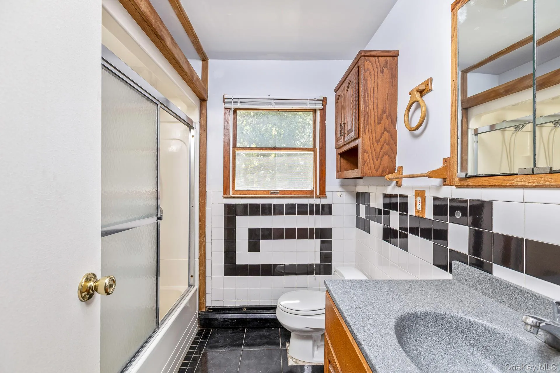 Bathroom featuring a shower-tub combination with sliding glass doors, a window with wood trim, a wood-finish vanity with an integrated sink, and black and white tiled walls Bathroom featuring a shower-tub combination with sliding glass doors, a window with wood trim, a wood-finish vanity with an integrated sink, and black and white tiled walls