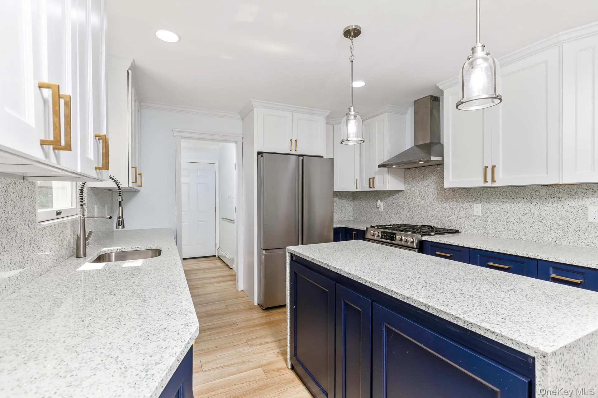 White upper cabinetry with gold-finish hardware, blue lower cabinetry with coordinating hardware, light-toned wood-finish flooring, white speckled countertops, and stainless steel appliances White upper cabinetry with gold-finish hardware, blue lower cabinetry with coordinating hardware, light-toned wood-finish flooring, white speckled countertops, and stainless steel appliances
