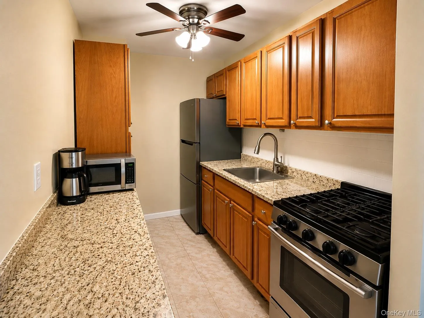 Kitchen featuring wood cabinetry, stainless steel appliances, granite-style countertops, tile flooring, and a ceiling fan Kitchen featuring wood cabinetry, stainless steel appliances, granite-style countertops, tile flooring, and a ceiling fan