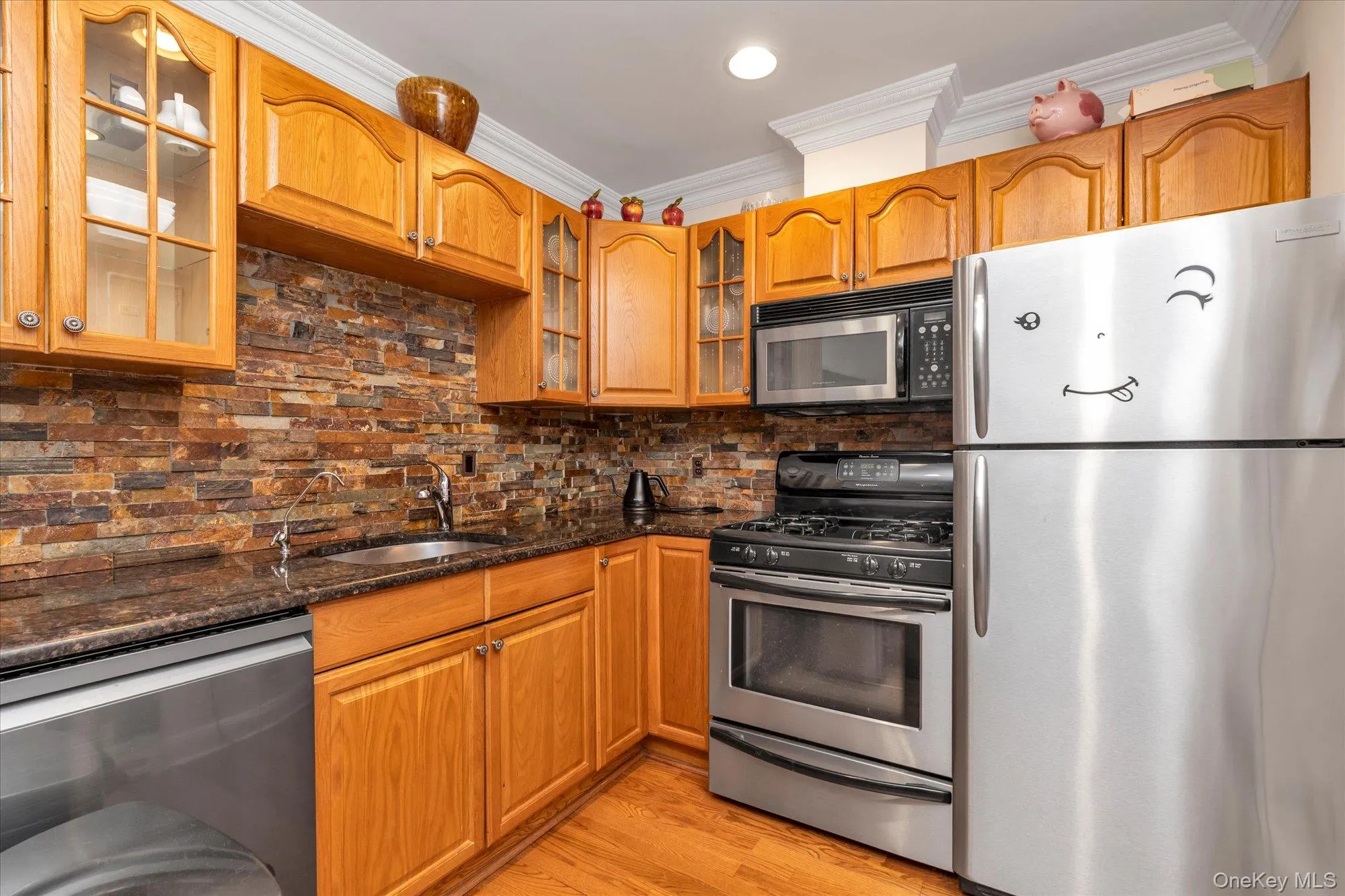 Kitchen featuring wood cabinetry, dark speckled countertops, stone-look tile backsplash, stainless steel appliances, and wood-finish flooring Kitchen featuring wood cabinetry, dark speckled countertops, stone-look tile backsplash, stainless steel appliances, and wood-finish flooring
