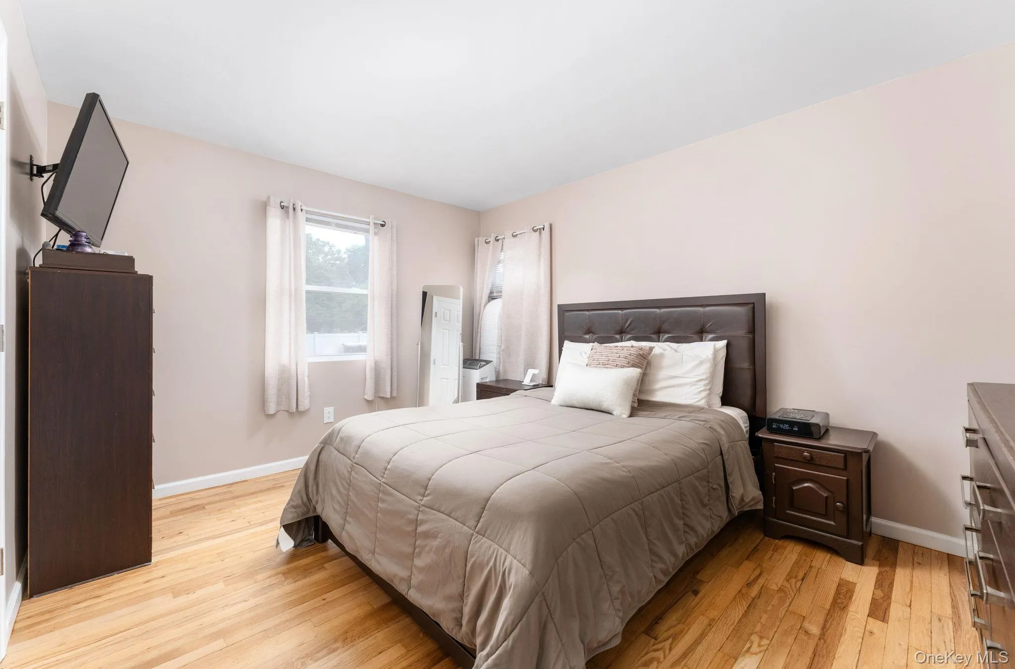Bedroom featuring light wood-finish flooring, a window with white curtains, a tufted dark-toned headboard, and light-toned wall paint Bedroom featuring light wood-finish flooring, a window with white curtains, a tufted dark-toned headboard, and light-toned wall paint