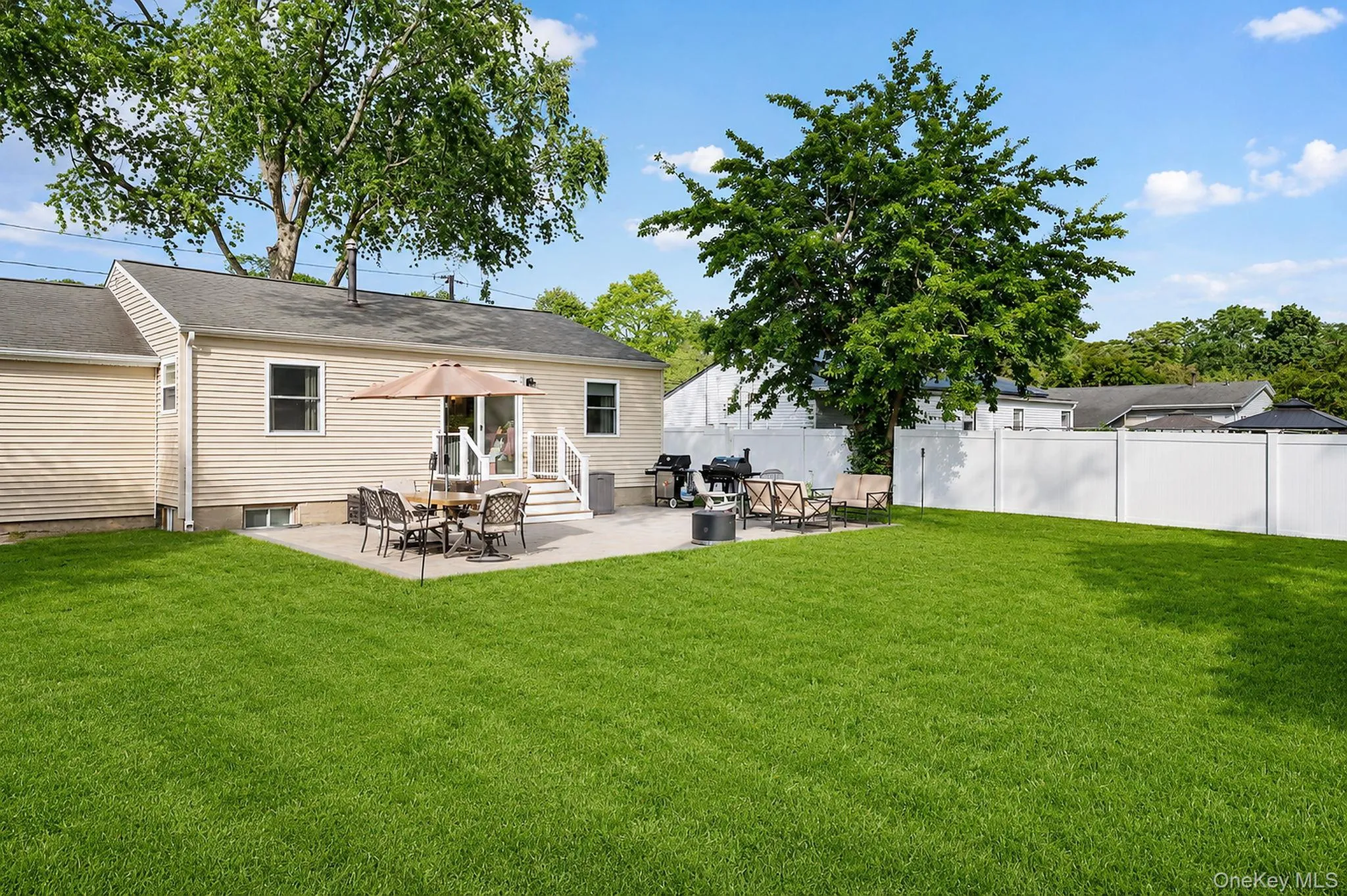 Expansive lawn area featuring a concrete patio, white vinyl fencing, and mature trees Expansive lawn area featuring a concrete patio, white vinyl fencing, and mature trees