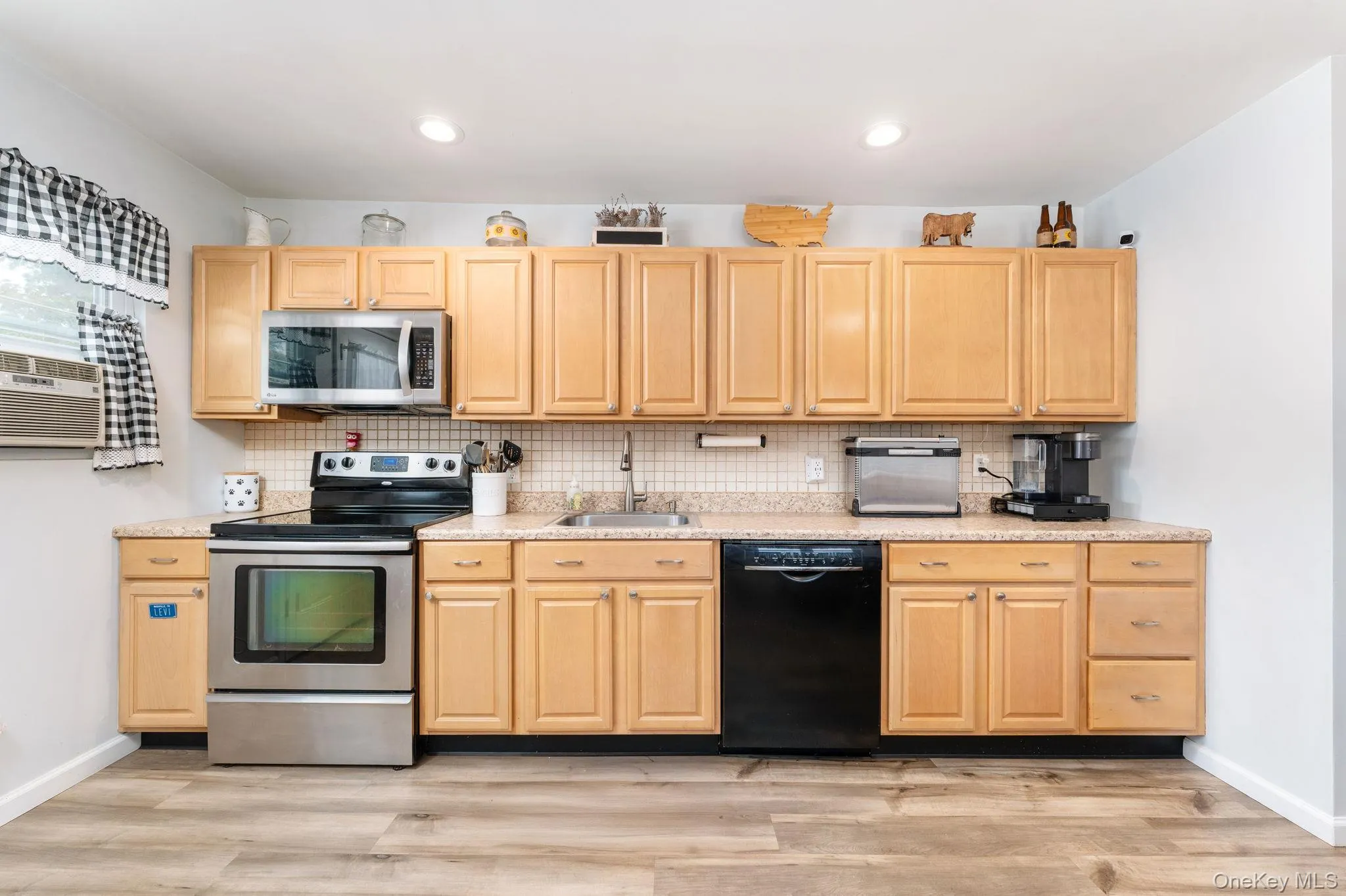 Kitchen featuring wood-finish flooring, light wood cabinetry, tile backsplash, stainless steel appliances, and a black dishwasher Kitchen featuring wood-finish flooring, light wood cabinetry, tile backsplash, stainless steel appliances, and a black dishwasher