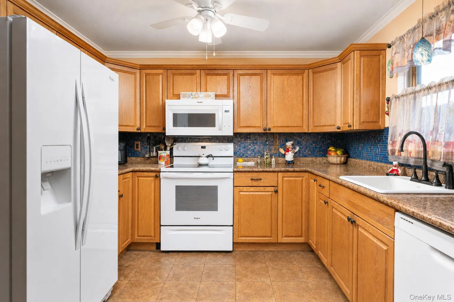 Kitchen featuring wood cabinetry, white appliances, a blue mosaic tile backsplash, and a white ceiling fan Kitchen featuring wood cabinetry, white appliances, a blue mosaic tile backsplash, and a white ceiling fan