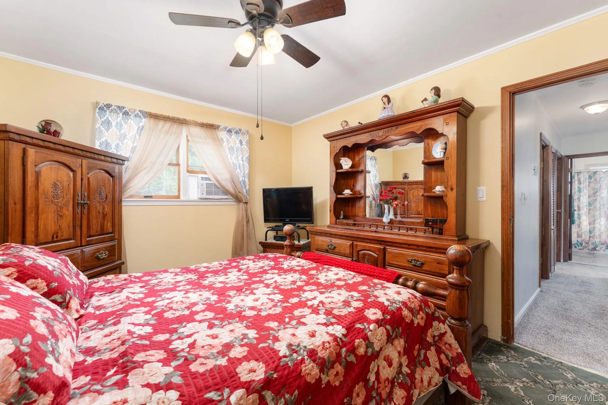 Bedroom featuring a ceiling fan with integrated lighting, a window with an installed air conditioning unit, and a doorway leading to a carpeted hallway Bedroom featuring a ceiling fan with integrated lighting, a window with an installed air conditioning unit, and a doorway leading to a carpeted hallway