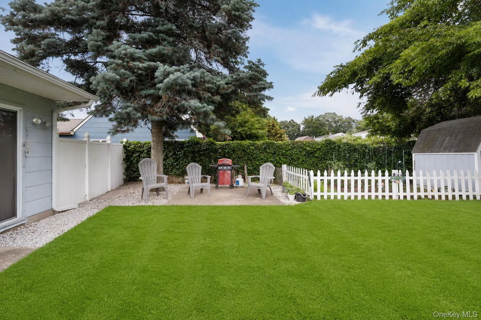 Spacious green lawn featuring a white picket fence, mature trees, a gravel pathway, and a patio area with a storage shed Spacious green lawn featuring a white picket fence, mature trees, a gravel pathway, and a patio area with a storage shed
