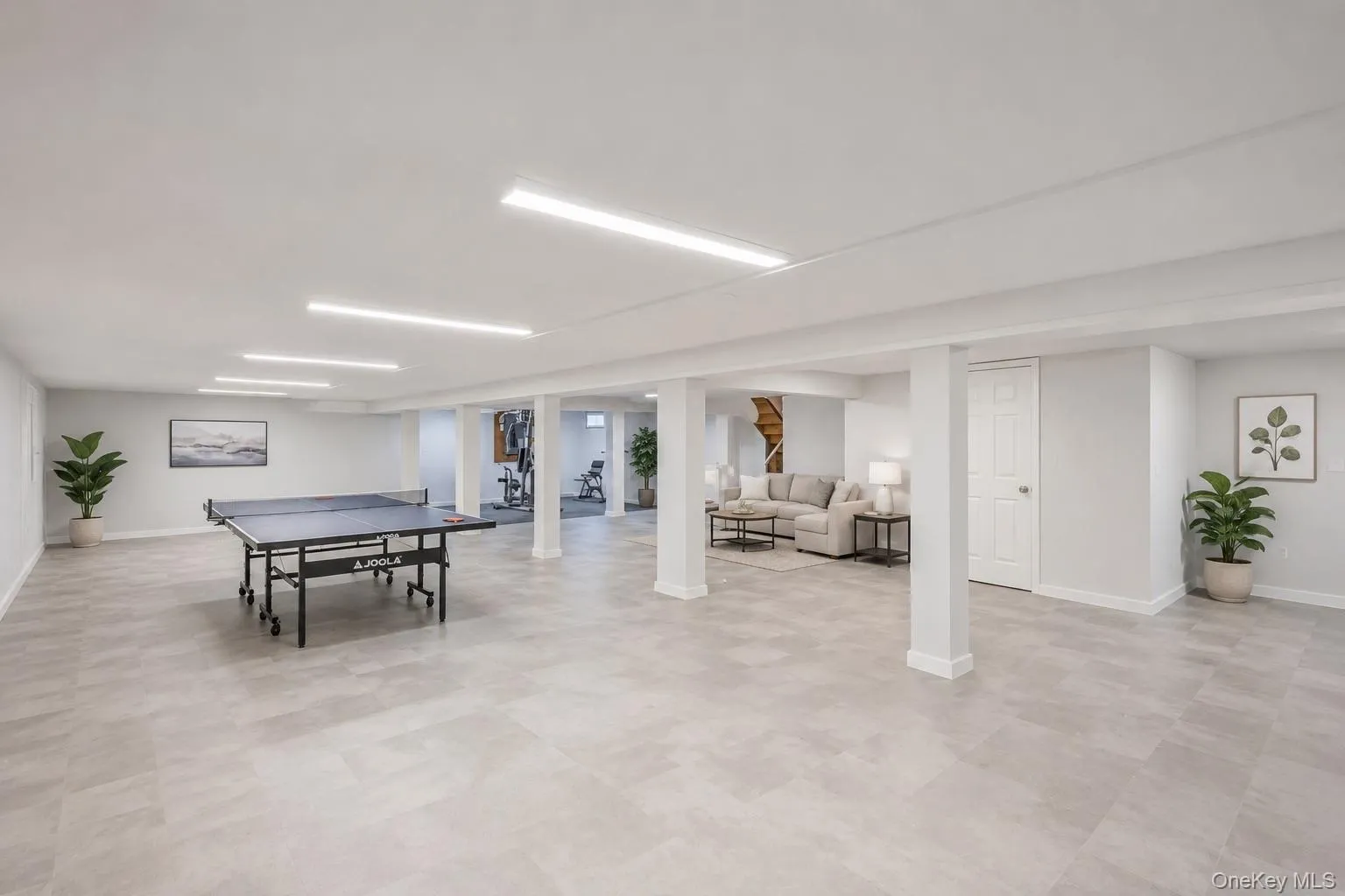 Spacious finished basement featuring recessed linear lighting, light gray floor tiles, painted support columns, a white door, and visible stairs Spacious finished basement featuring recessed linear lighting, light gray floor tiles, painted support columns, a white door, and visible stairs
